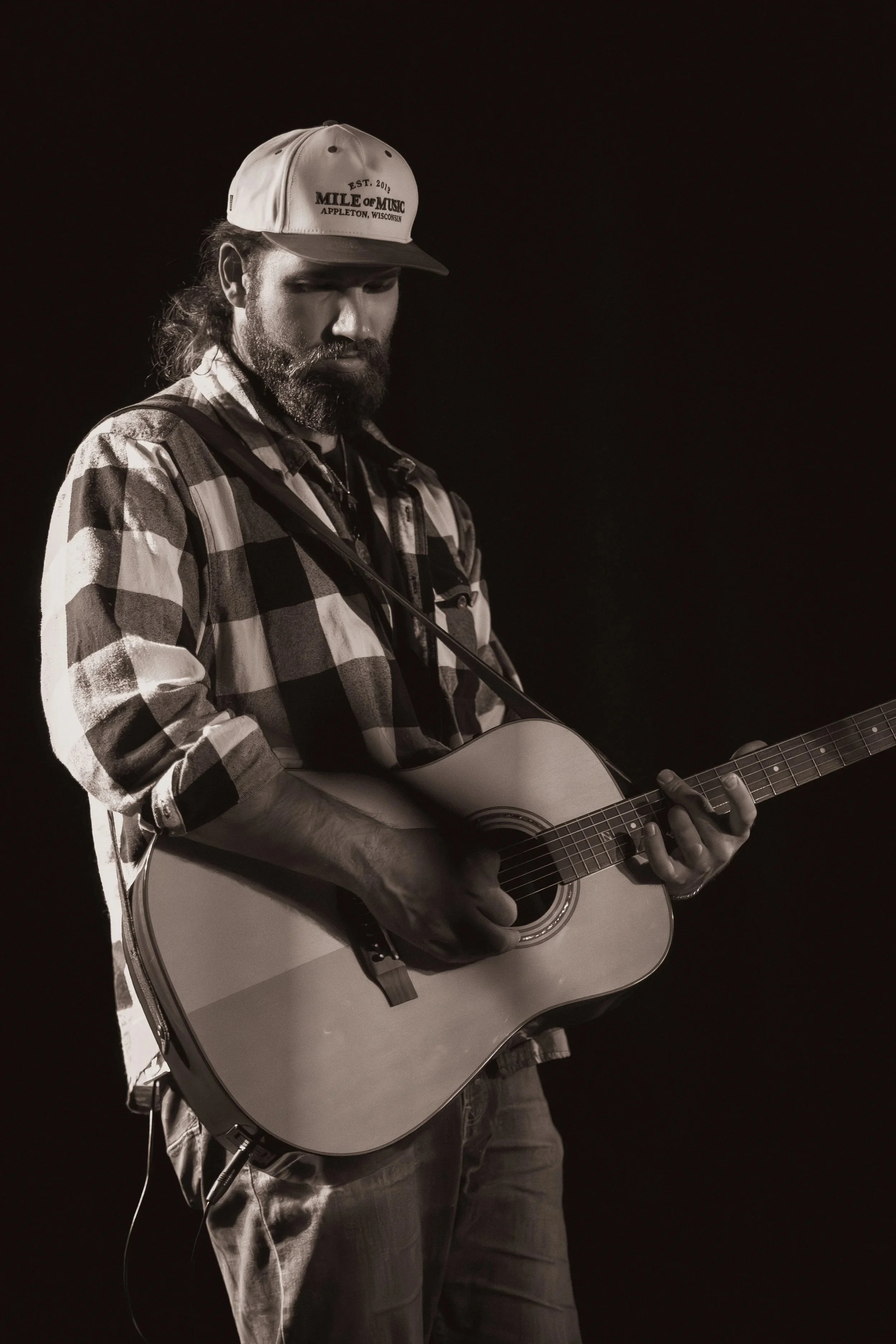 Black and white photo of a man with long hair and beard playing an acoustic guitar, wearing a checkered shirt and a baseball cap that says 'Mile of Music Appleton Wisconsin'.