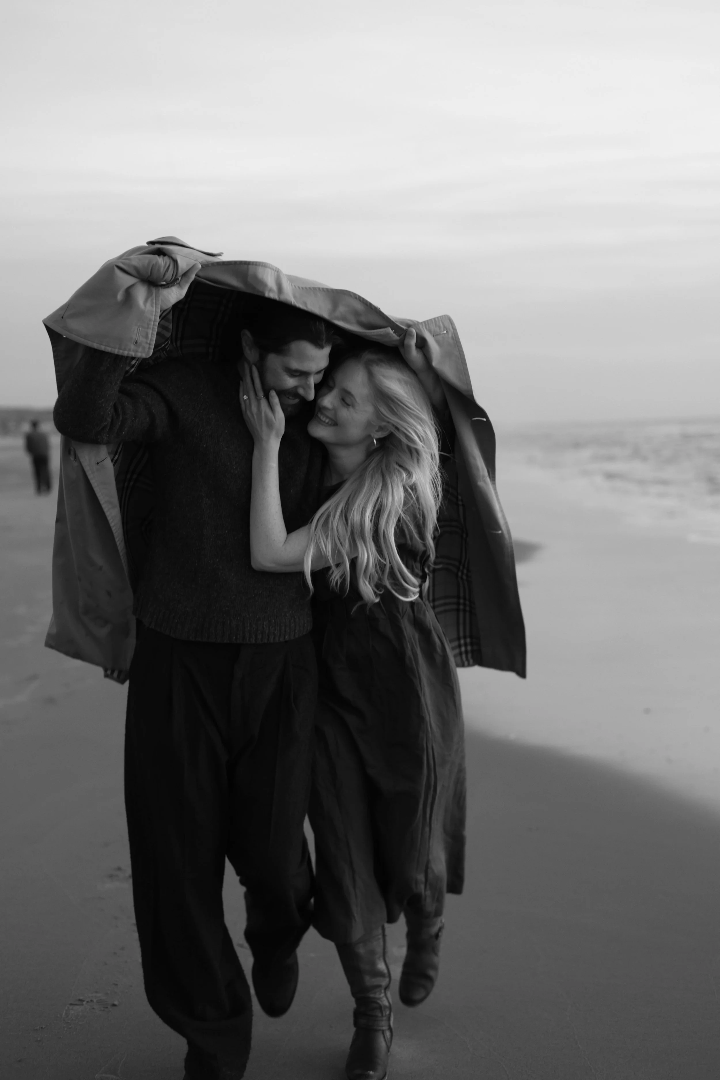 A black-and-white photo of a smiling couple walking on the beach, with the man holding a coat over both of their heads to shield them from the wind or weather.