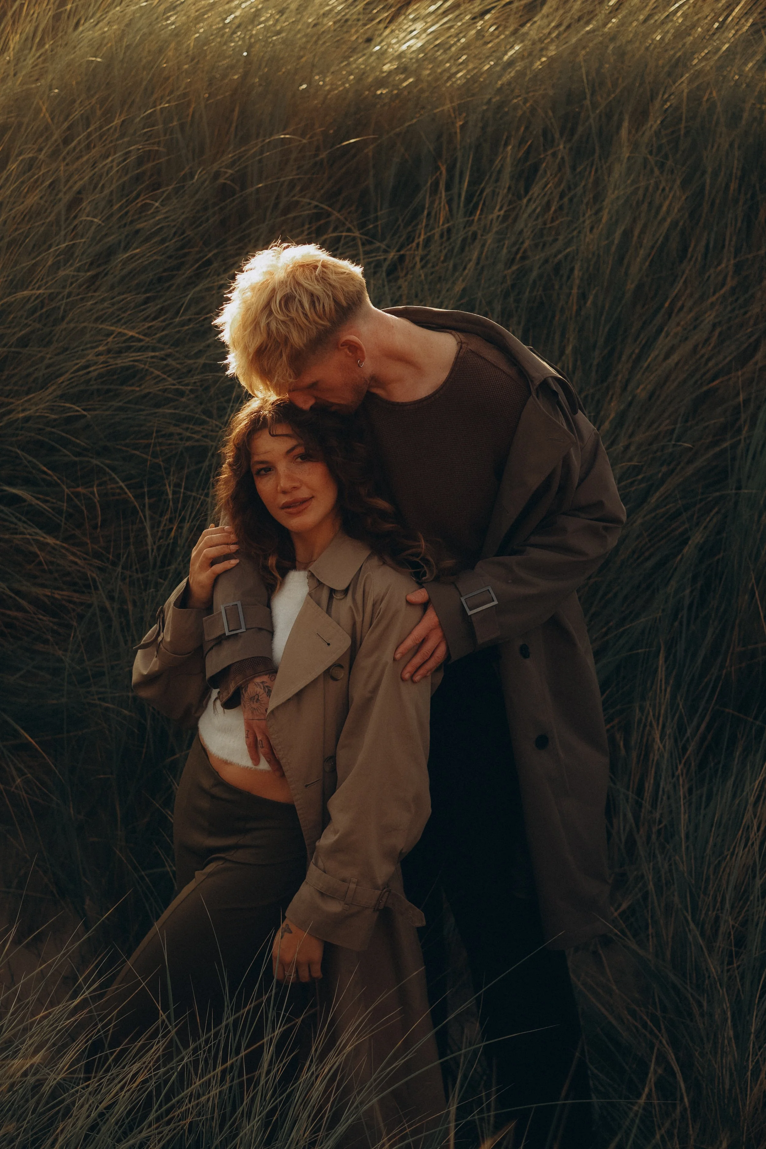 A man with blonde hair and a woman with dark hair posing amidst tall grasses, with the man leaning over the woman and touching her shoulder and the woman looking at the camera.