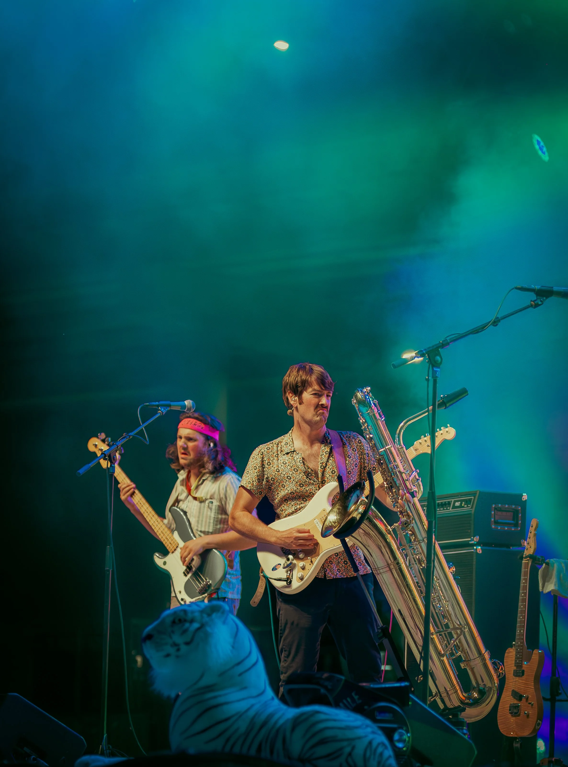 Musicians performing on stage with colorful lighting, surrounded by musical instruments including guitars, a saxophone, and microphones.