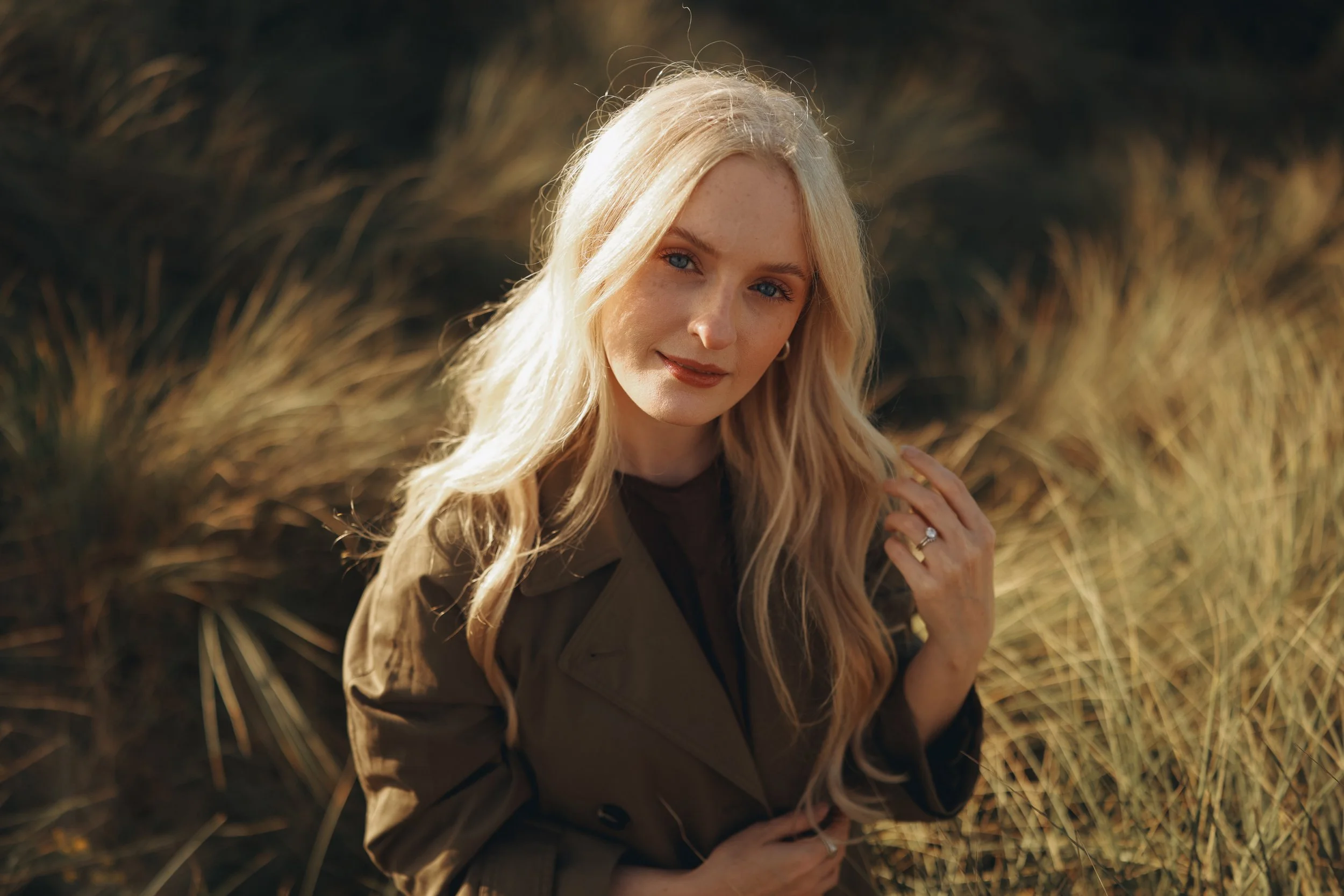 A blonde woman with blue eyes, wearing makeup, earrings, and a brown coat, smiling softly, standing outdoors among tall grass in warm sunlight.