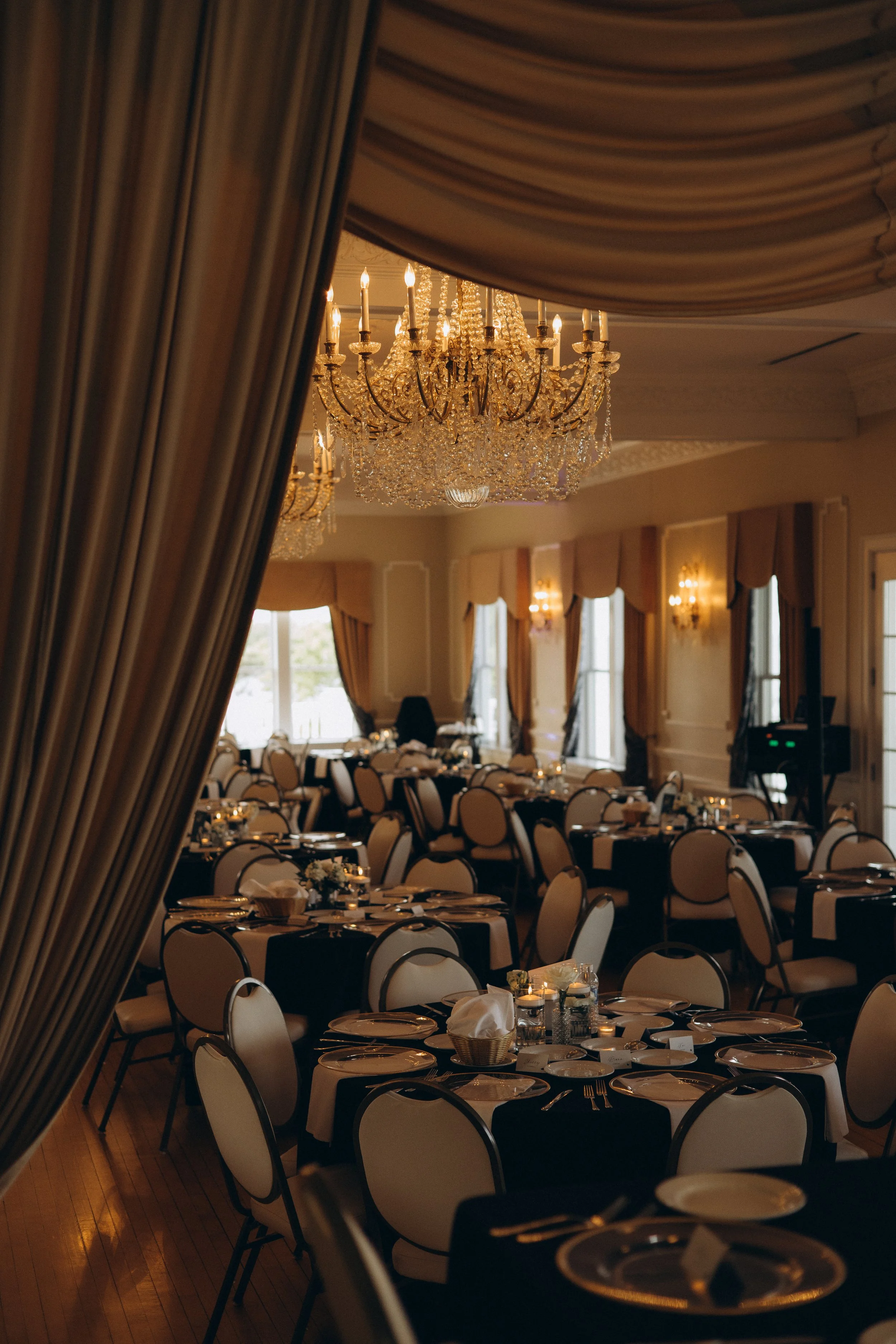 Elegant dining room with round tables set with plates, napkins, and utensils, decorated with candles, and large crystal chandeliers hanging from the ceiling amid draped curtains and windows.