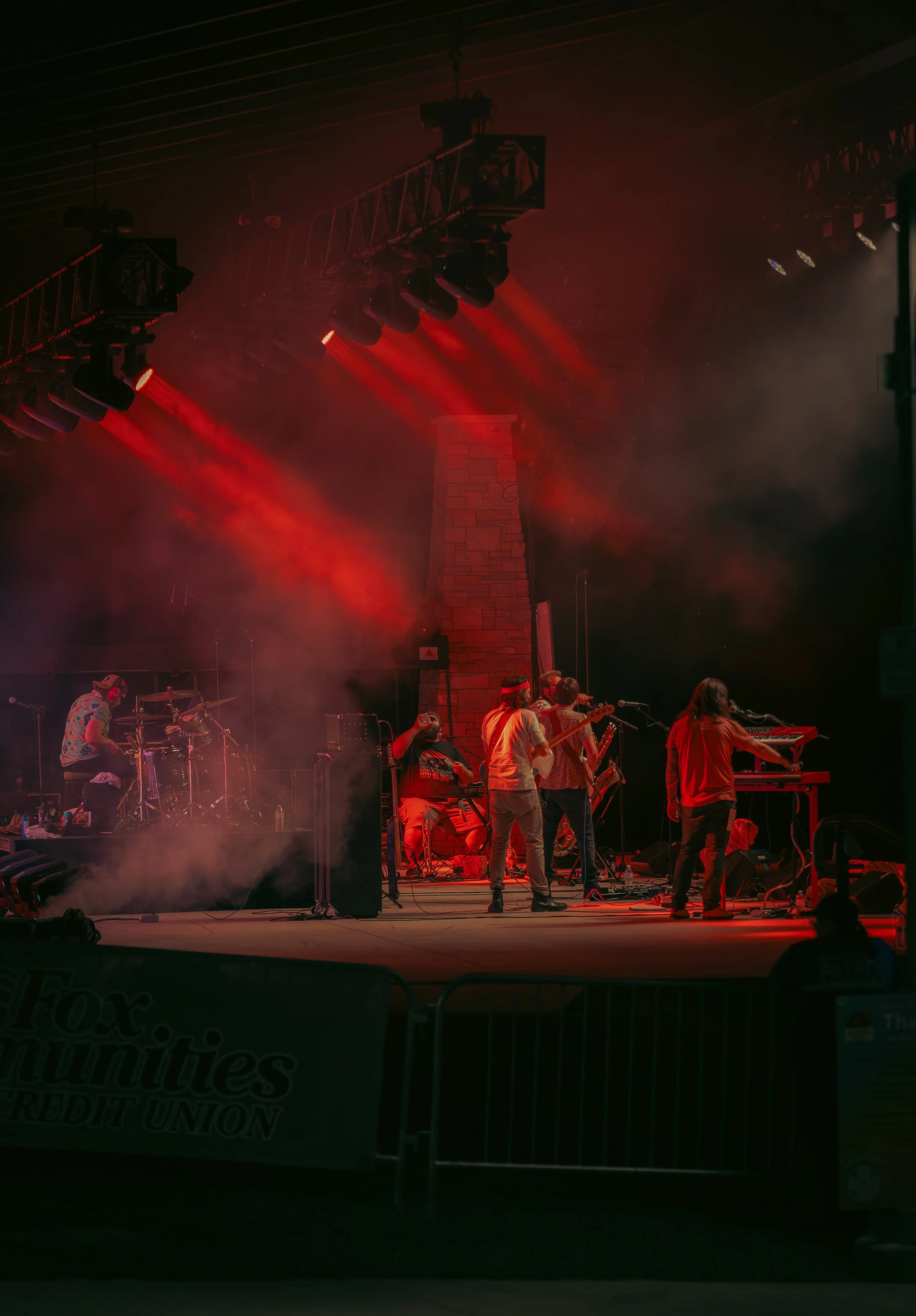 A live music performance at night with musicians on stage, red stage lights, and a crowd in front of the stage.