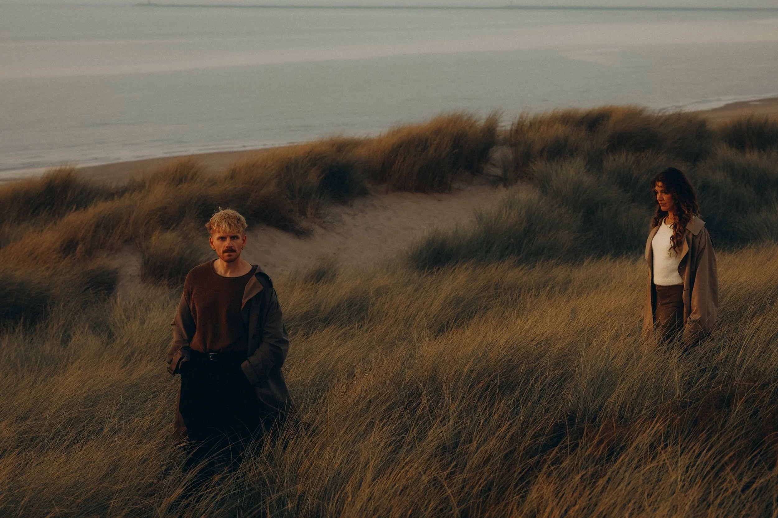 Two individuals walking through grassy dunes near the ocean during sunset or dusk.