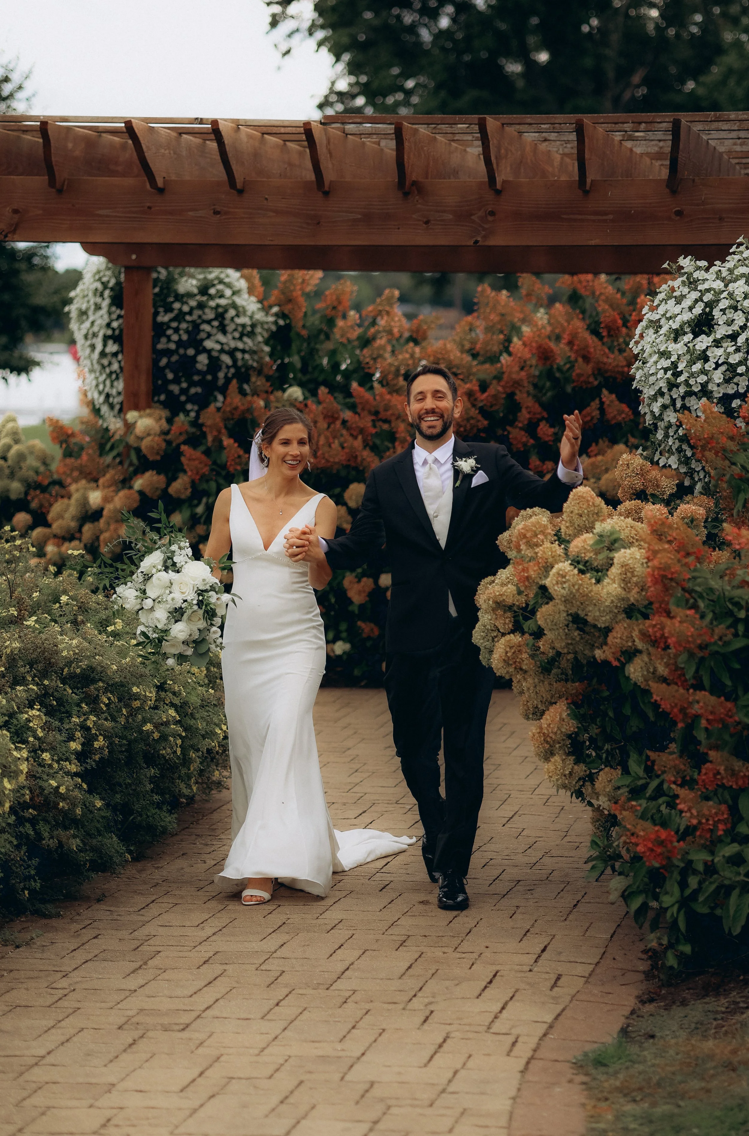A newlywed couple walking happily together down a brick path outdoors, surrounded by vibrant flowers and greenery, under an archway.