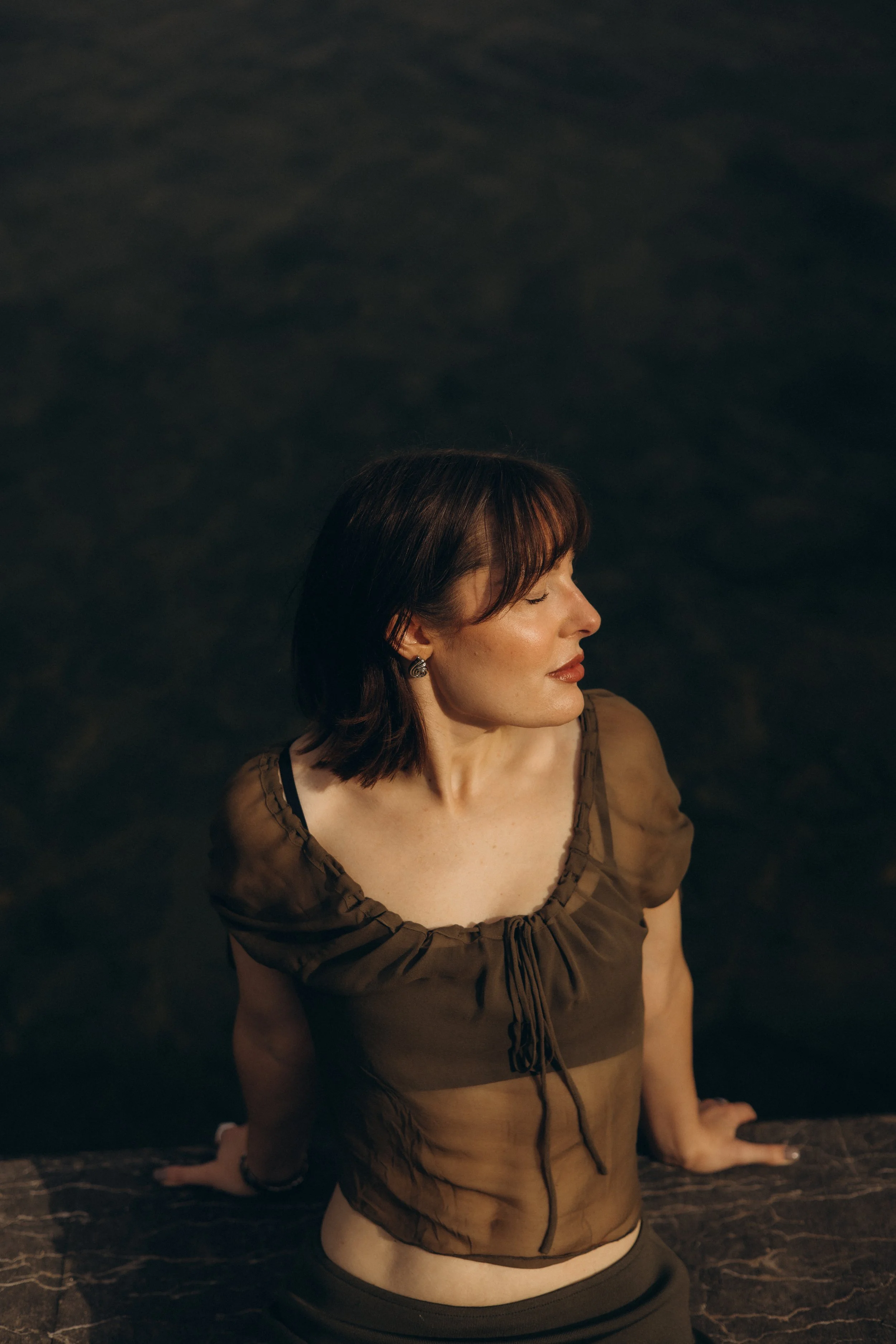A woman with short brown hair and earrings is sitting on a dark marble surface with her eyes closed and head tilted slightly to the side.