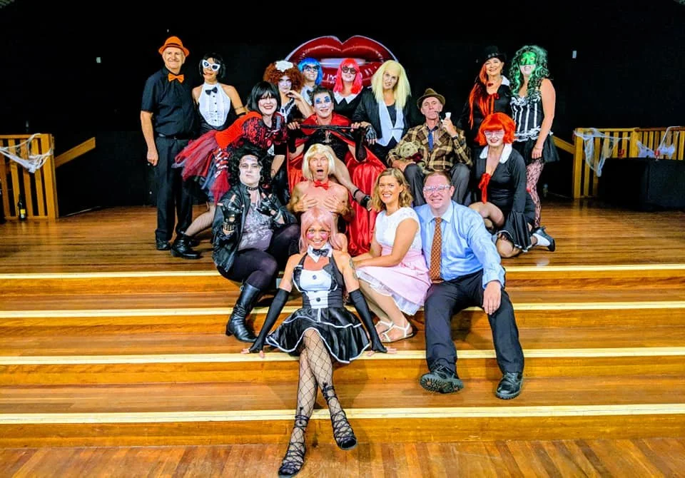 Group of people in costumes on a stage with a wooden floor, some sitting and some standing, with bright makeup and accessories, in front of a backdrop featuring red lips.