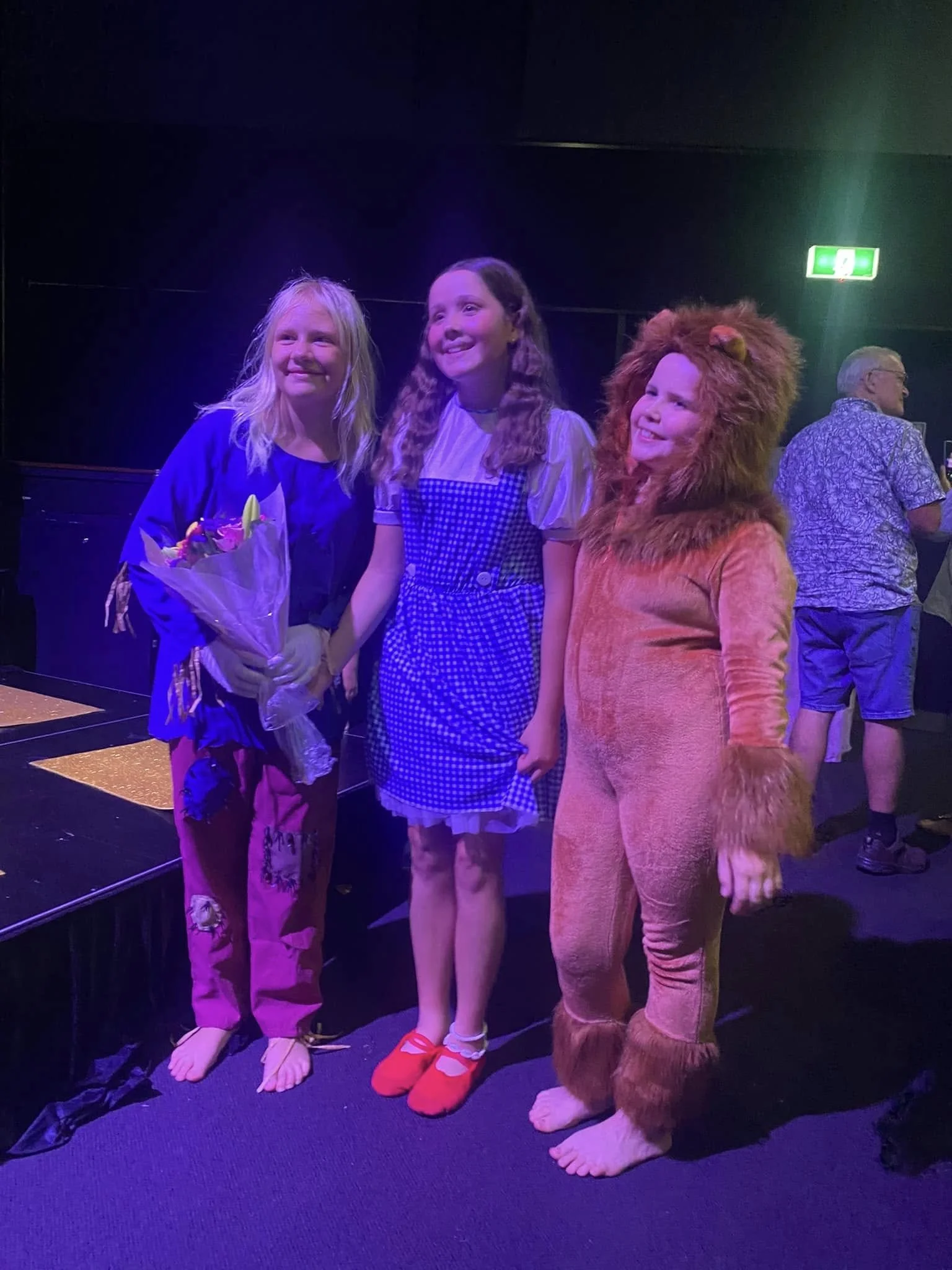 Three children on stage, one dressed as a lion, smiling and posing for a photo, with a man in the background.