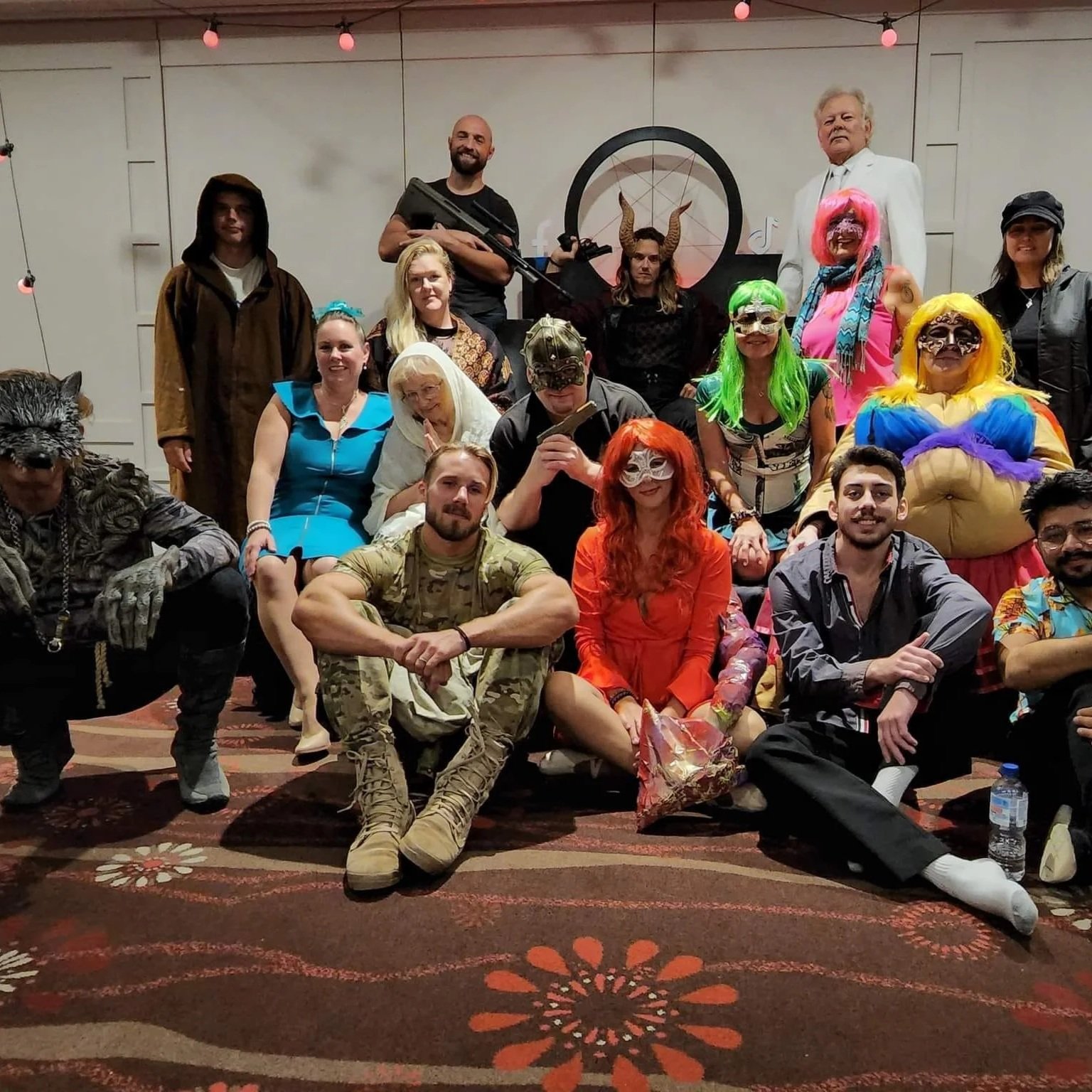 Group of people in costumes at a party or cosplay event, some wearing masks, wigs, and themed outfits, with a man holding a prop gun in the back.