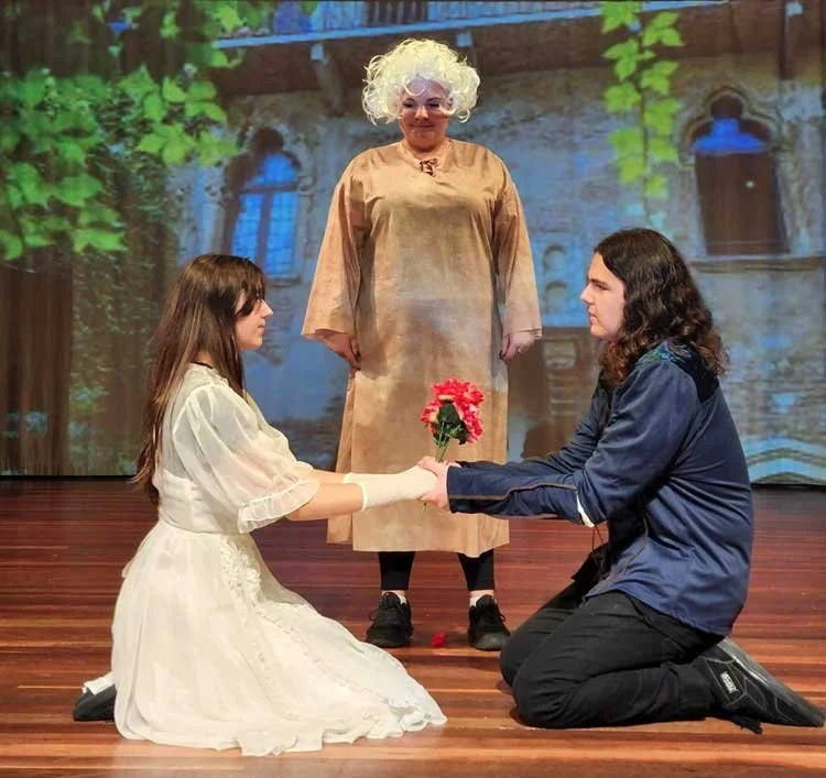 A theatrical scene with two young women kneeling on a wooden stage, facing each other and holding hands. The woman on the left is dressed in a white costume, and the woman on the right wears a dark blue jacket. The woman on the left is offering a bouquet of red flowers to the woman on the right. A person stands behind them, observing, dressed in a beige costume with curly white hair, set against a backdrop resembling an old building with green foliage.