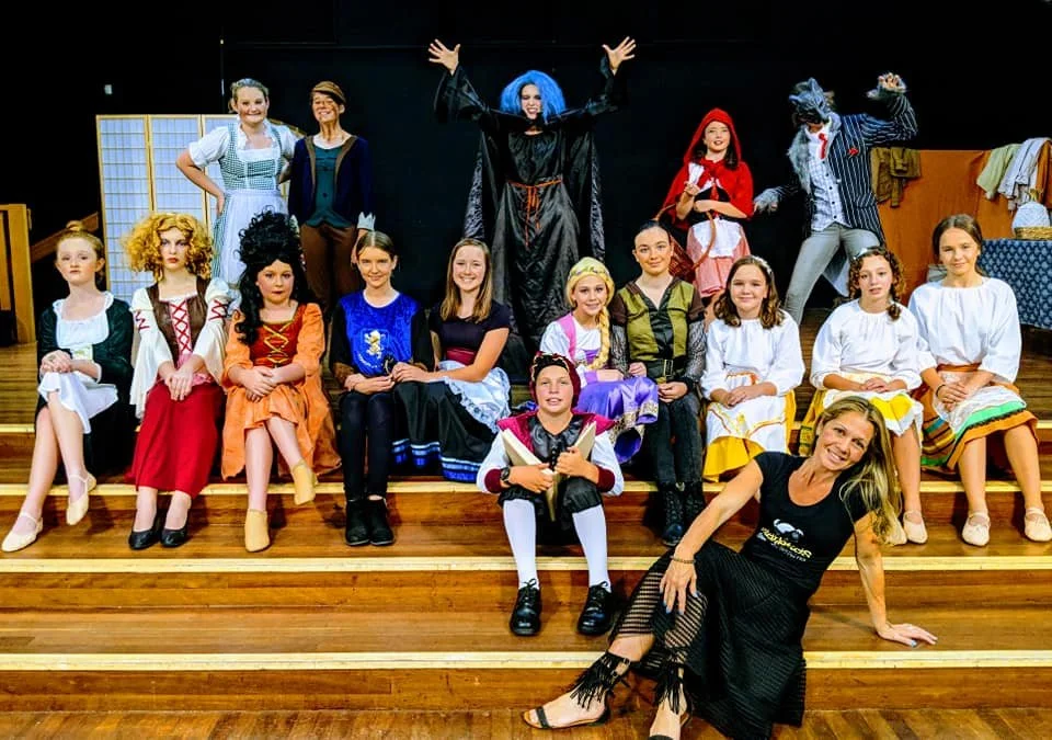 Group of children and adults in costumes on stage with wooden stairs, some characters in fairy tale or storybook costumes, and stage props.