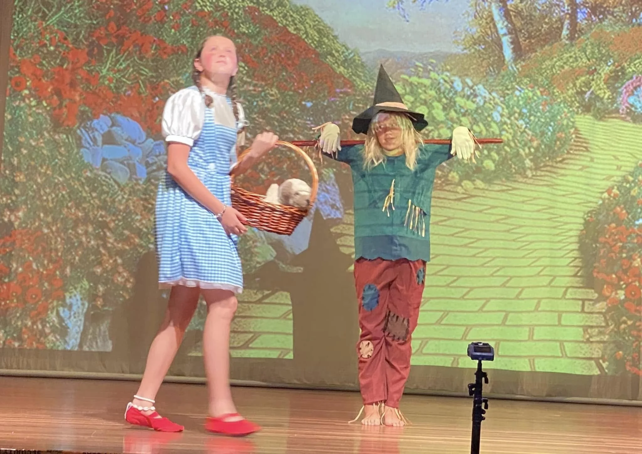 Two children on stage dressed as characters from 'The Wizard of Oz'; the girl as Dorothy holding a basket with a puppy, and the boy as the Scarecrow wearing a hat and patchy clothing, with a tree and garden background projected behind them.