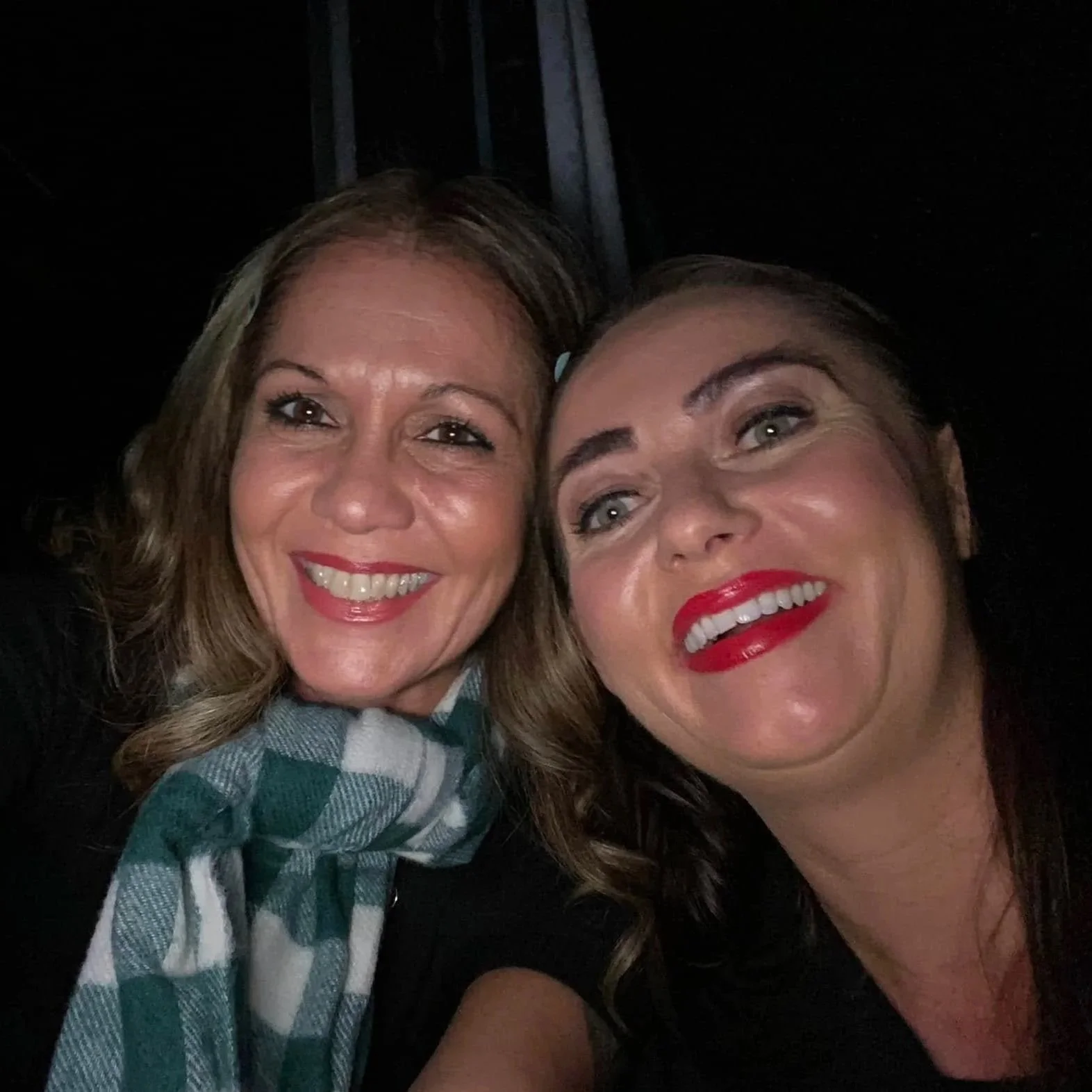 Two women smiling closely for a selfie, one with a scarf and the other with bright red lipstick.
