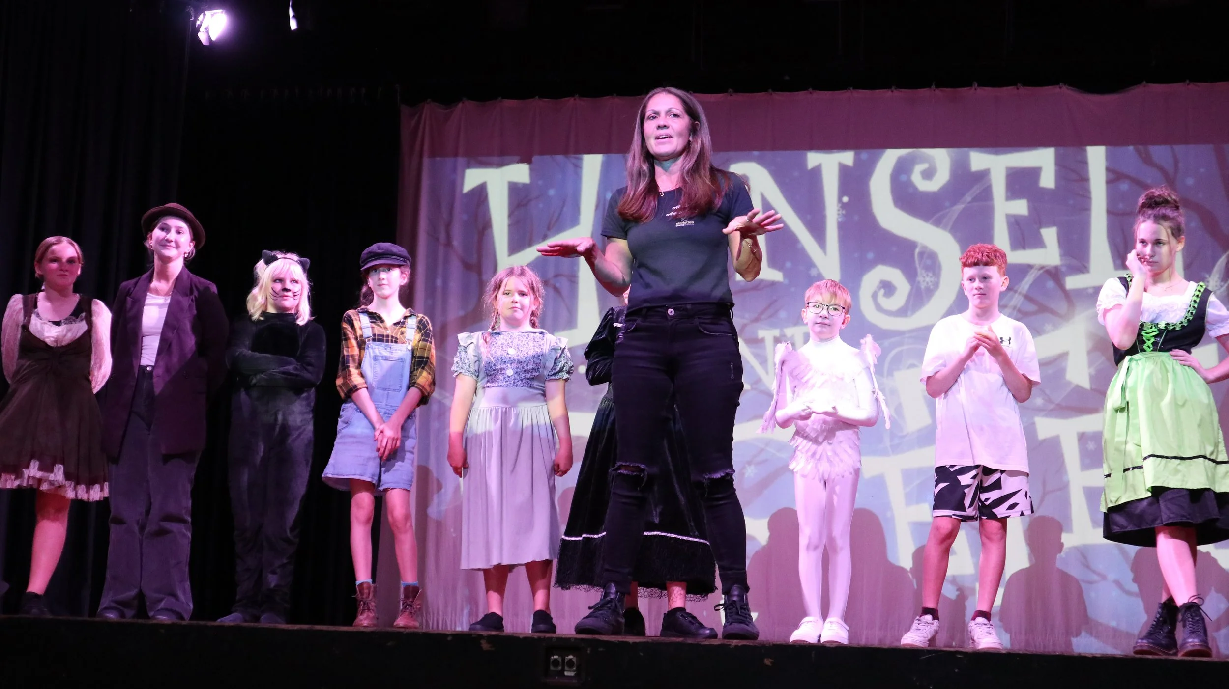 A woman standing on stage with children dressed in various costumes, possibly during a theater performance or rehearsal, with a backdrop that reads 'TINSLE' in colorful letters.