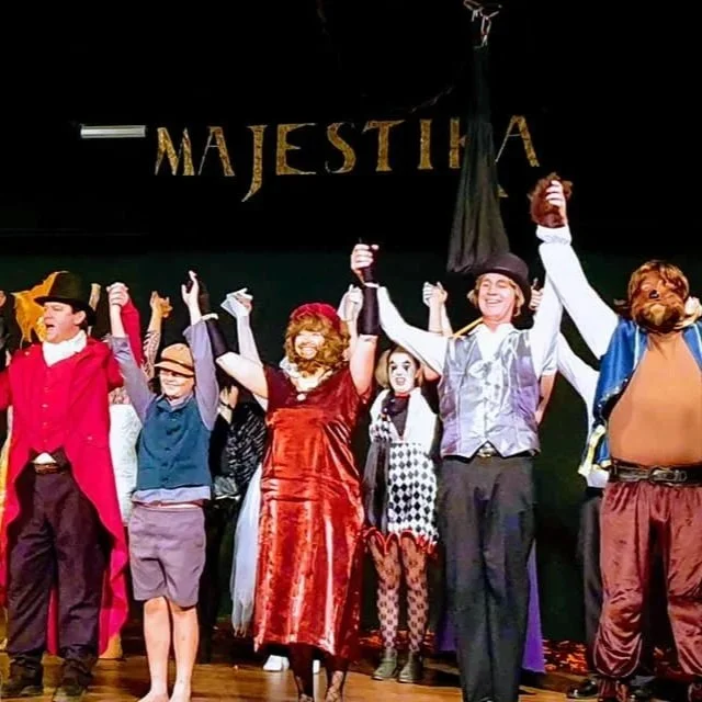 The cast of 'Majestic' on stage, dressed in colorful costumes, holding hands and taking a bow at the end of a theatrical performance.