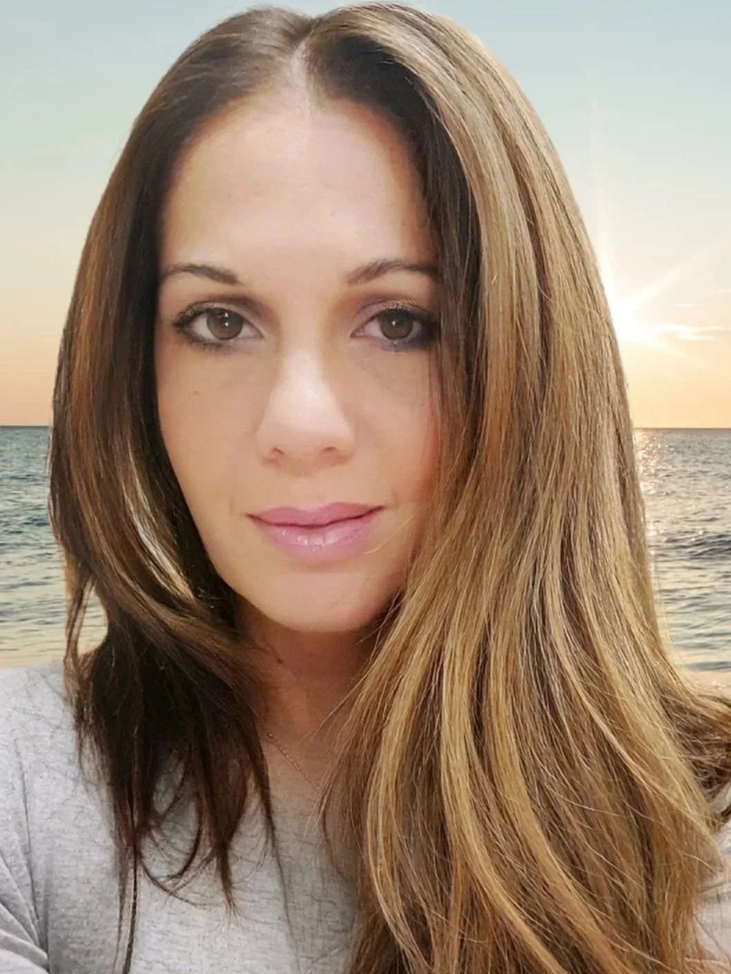 A woman with long chestnut hair and brown eyes stands in front of a sunset over the ocean.