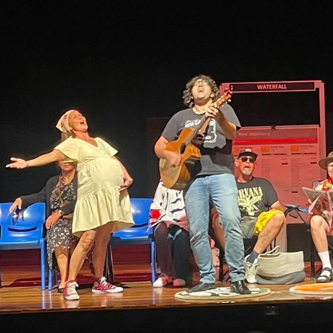 The image shows a group of people on stage, with one person standing and playing guitar, and others sitting on chairs behind them. The standing person appears to be singing passionately, while one woman in a yellow dress is gesturing with her arms.