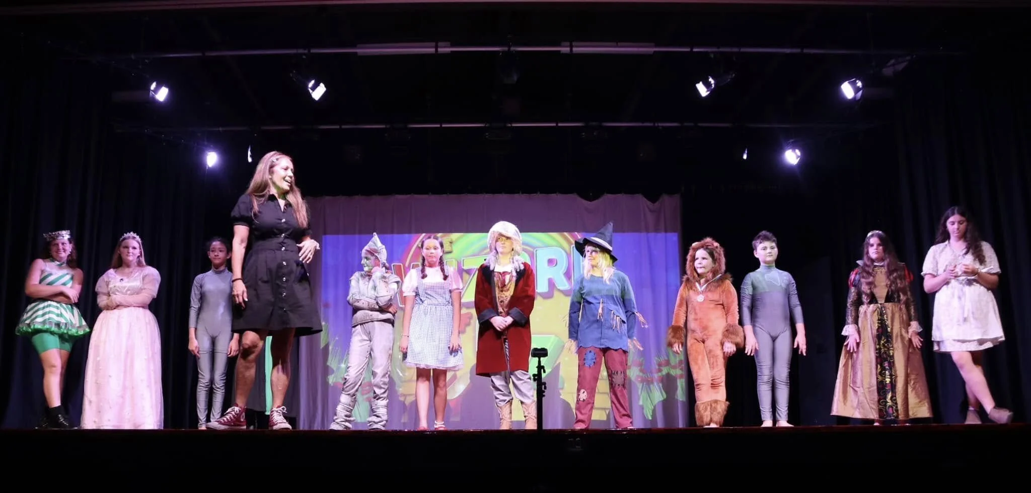 Children dressed in costumes standing on a stage with a woman in a black dress.