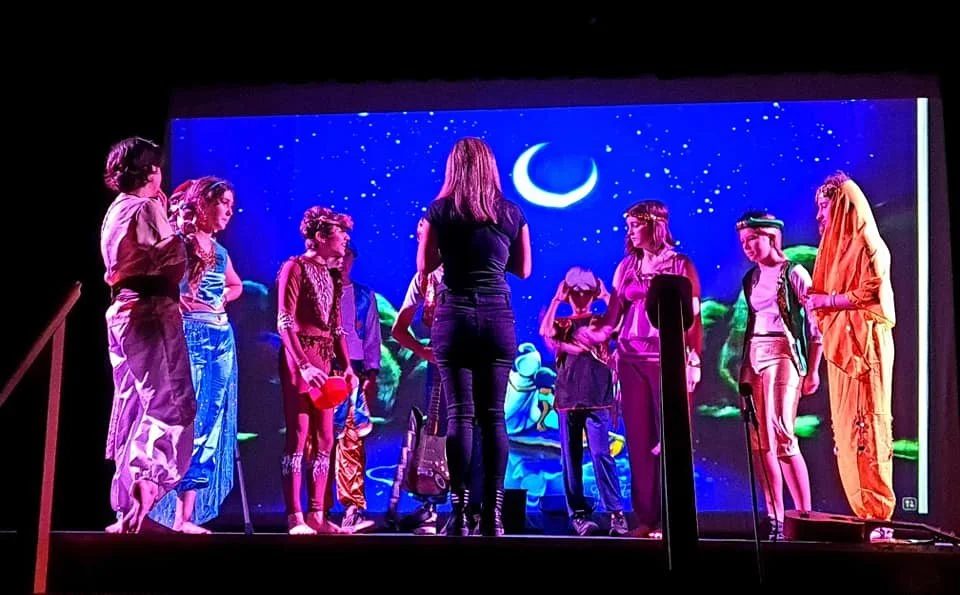 Children and a woman on stage during a performance with a colorful background of the night sky and moon.