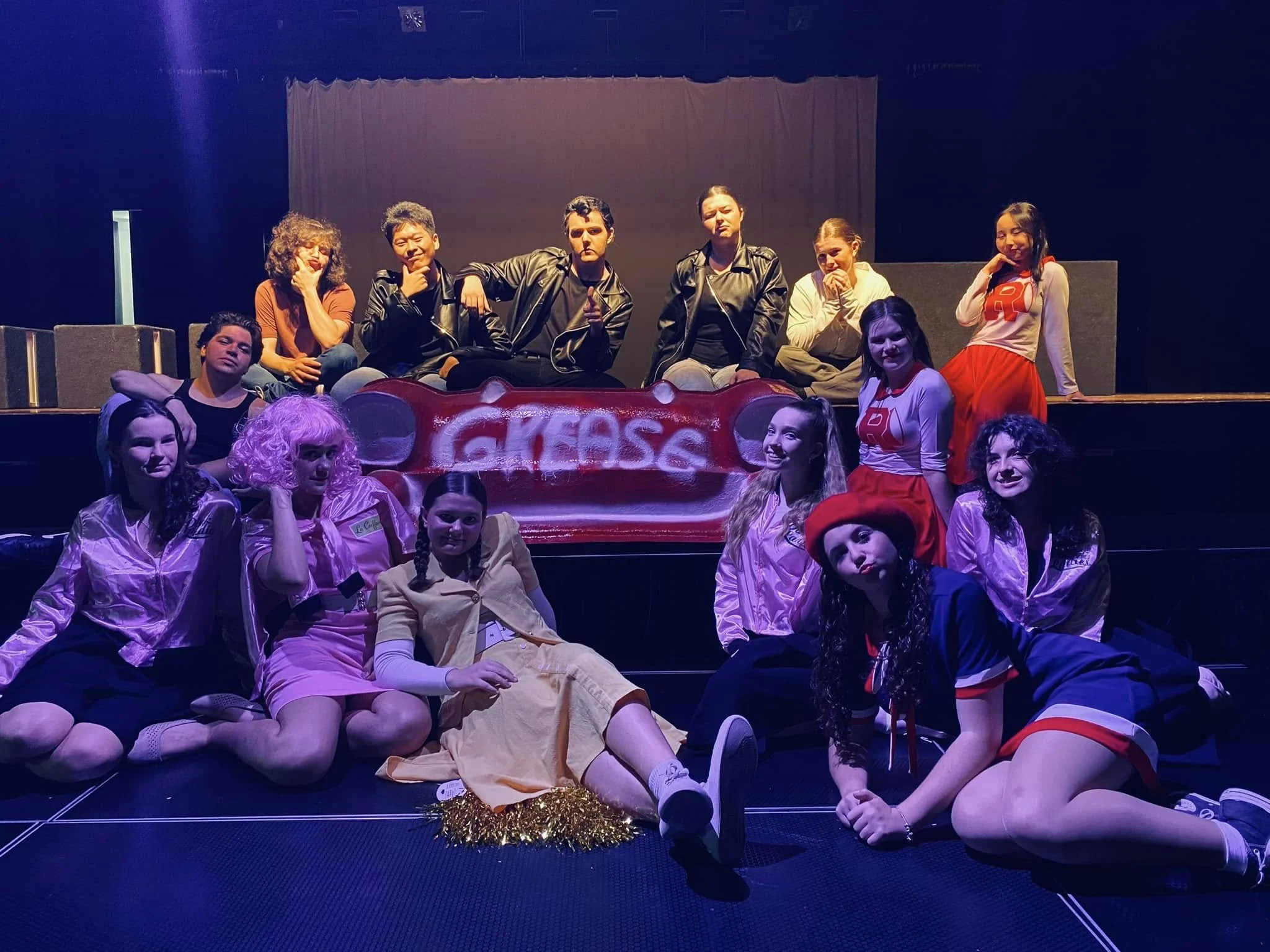A group of young people, mostly girls, posing on stage for a theatrical production or school play. They are dressed in costumes, with some sitting and some standing, in front of a red prop that reads "Glee" with mouse ears. The background is dark and
