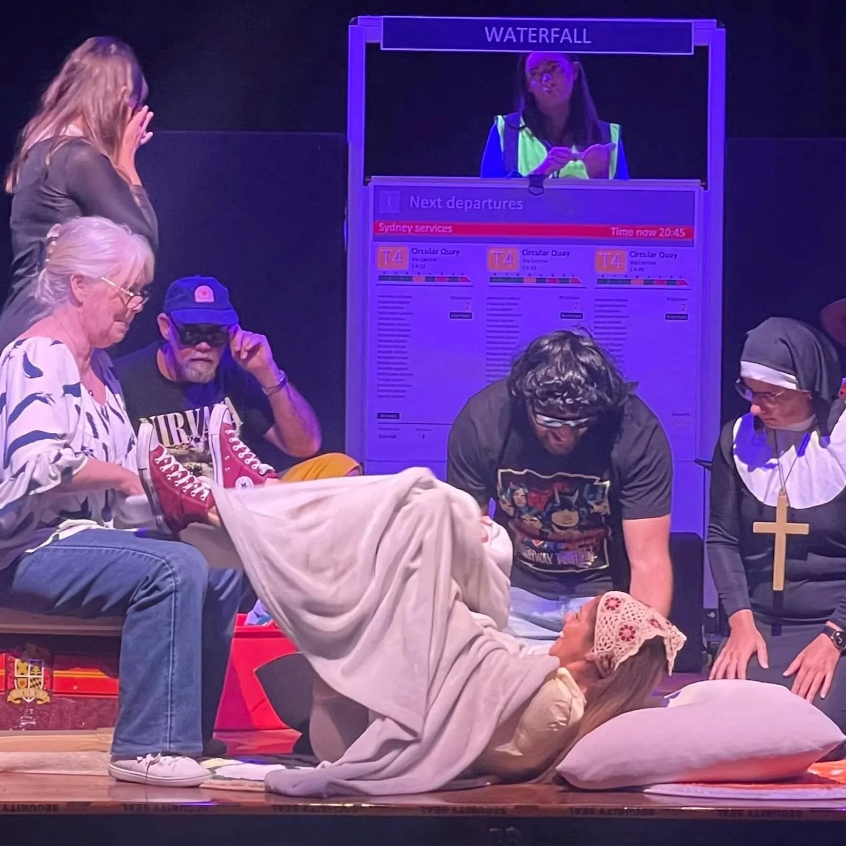 Stage scene resembling a religious reenactment with people surrounding a person lying on the ground, a woman dressed as Mary, others dressed casually, a person dressed as a nun, a woman with a colorful shirt, and a young girl with a head covering. There is a monitor displaying transportation schedules in the background.