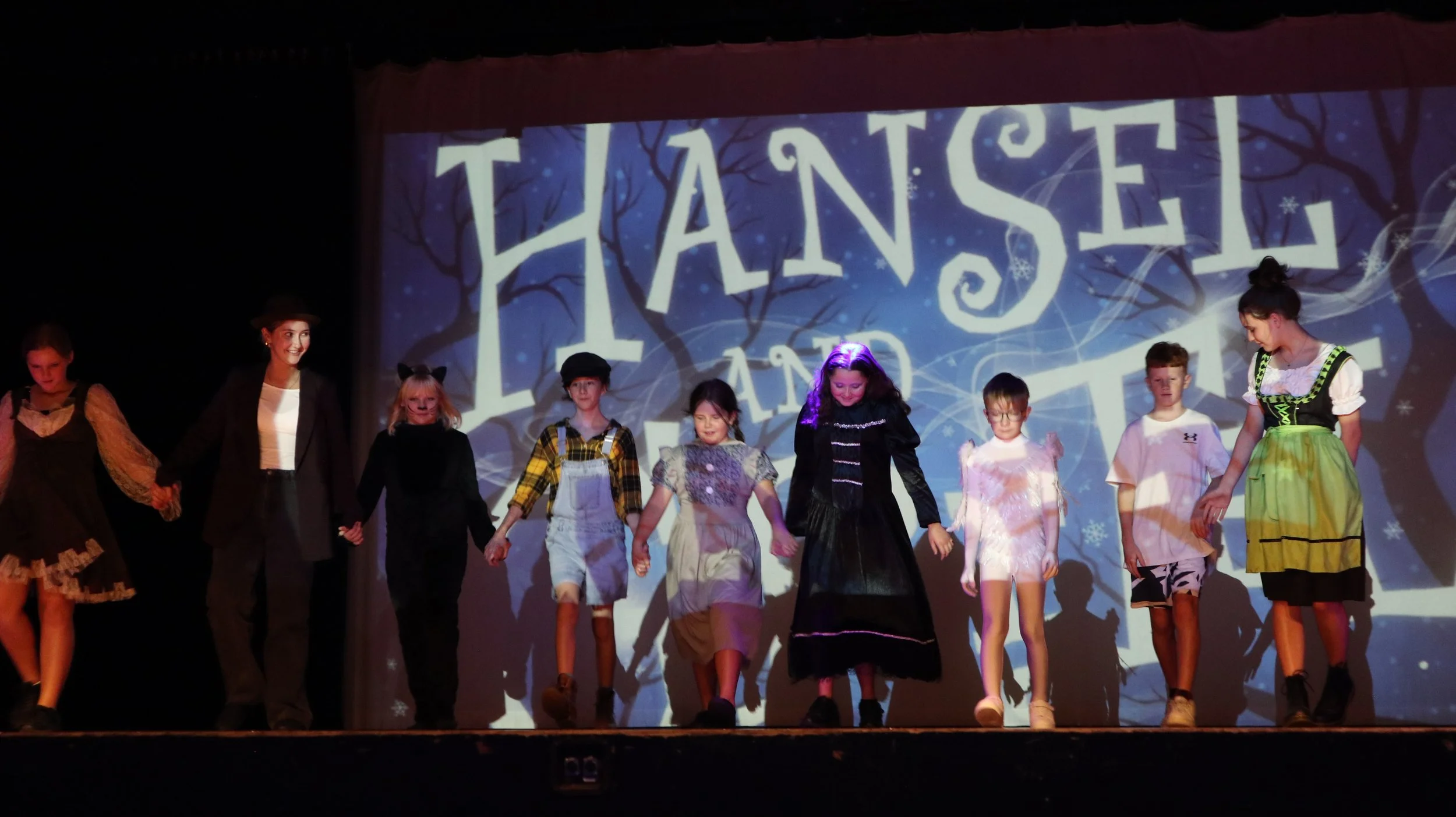 Children in costume holding hands on stage during a performance with a backdrop reading 'Hansel and Gretel'.