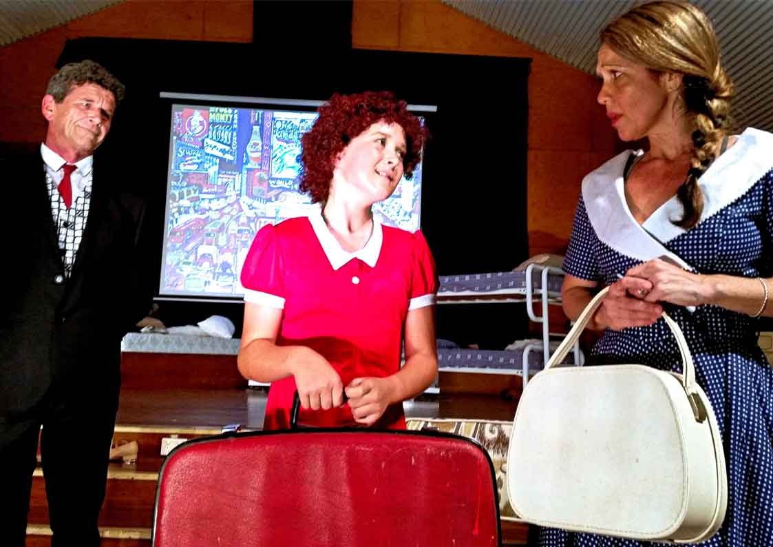 Three people indoors, with a colorful comic-style artwork on a screen in the background. The person in the middle, with curly red hair and a red dress, appears to be speaking or reacting, holding a red case. The person on the right, with blonde hair 