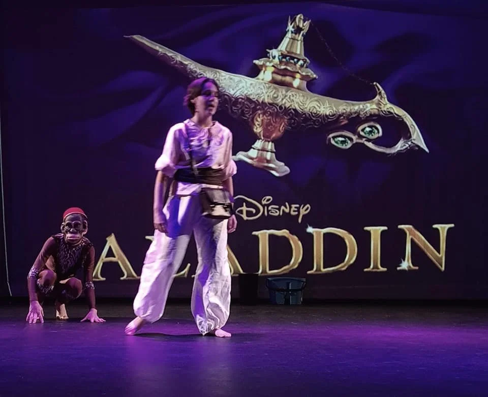 Two performers on stage, one standing and one crouching, in a theatrical performance of Disney's Aladdin. The background features a large screen displaying the Disney Aladdin logo with the magic carpet.