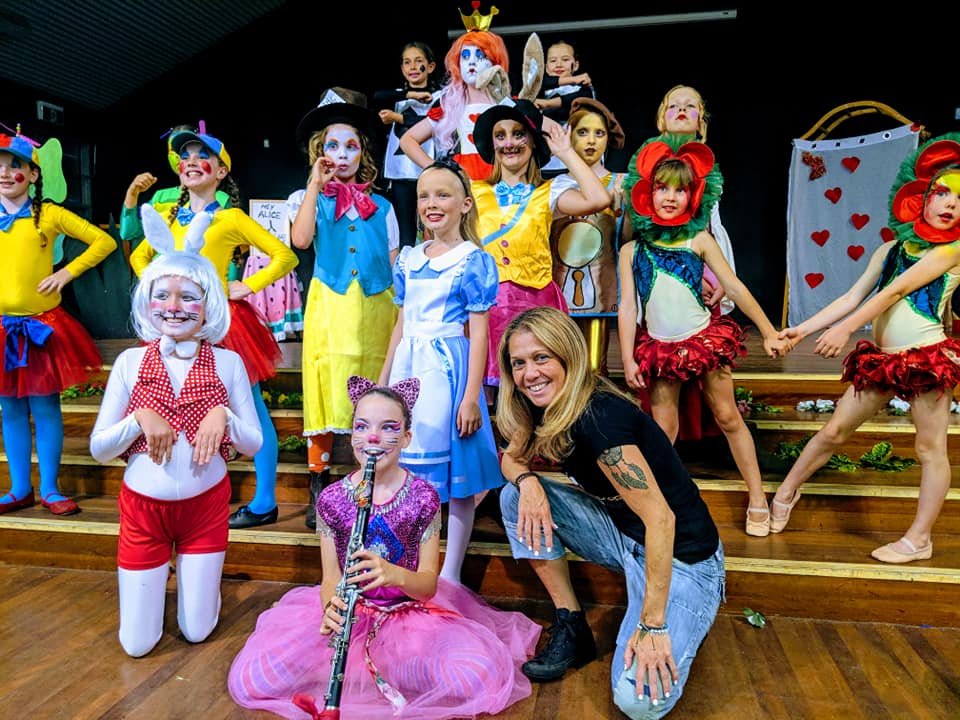 Children dressed in colorful costumes performing in a theatrical play with a woman in casual clothes, some children in animal costumes, and a clown with face paint.