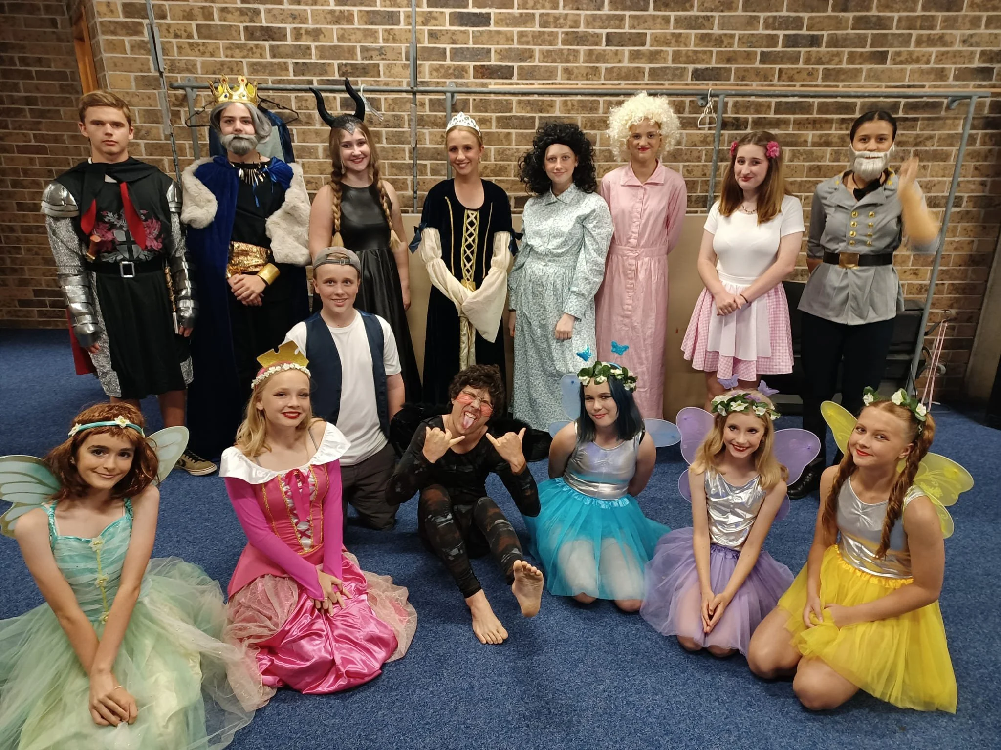 Group of children and an adult dressed in fairy tale and costume outfits, posing together in a room with brick wall background.