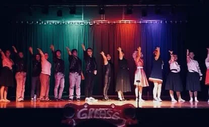 Group of performers standing on stage with colorful curtains in the background, under stage lights, participating in a dance or theater performance.