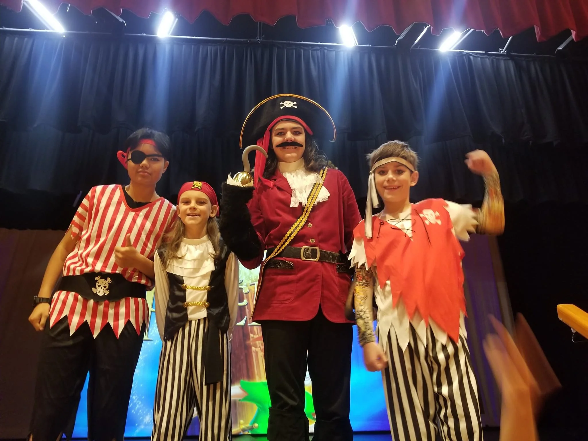 Children dressed in pirate and pirate-themed costumes on stage, posing for a group photo with a dark curtain backdrop and stage lights overhead.