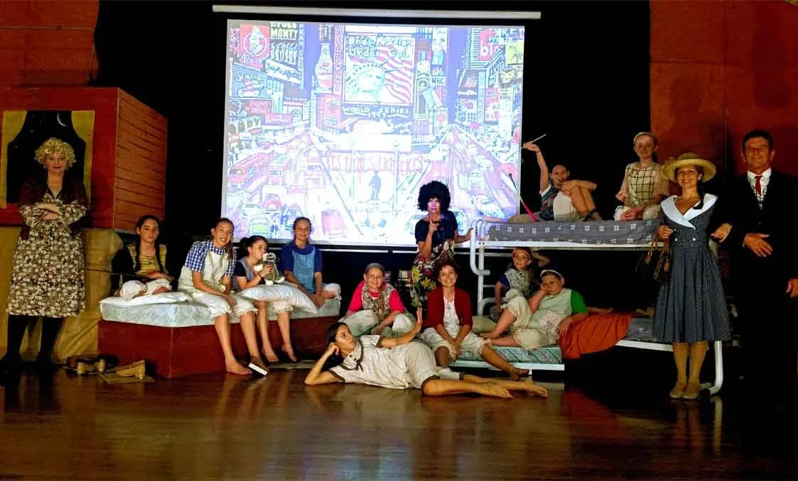 Children and adults participating in a theater play on stage with a backdrop of a cityscape and a projected movie scene.