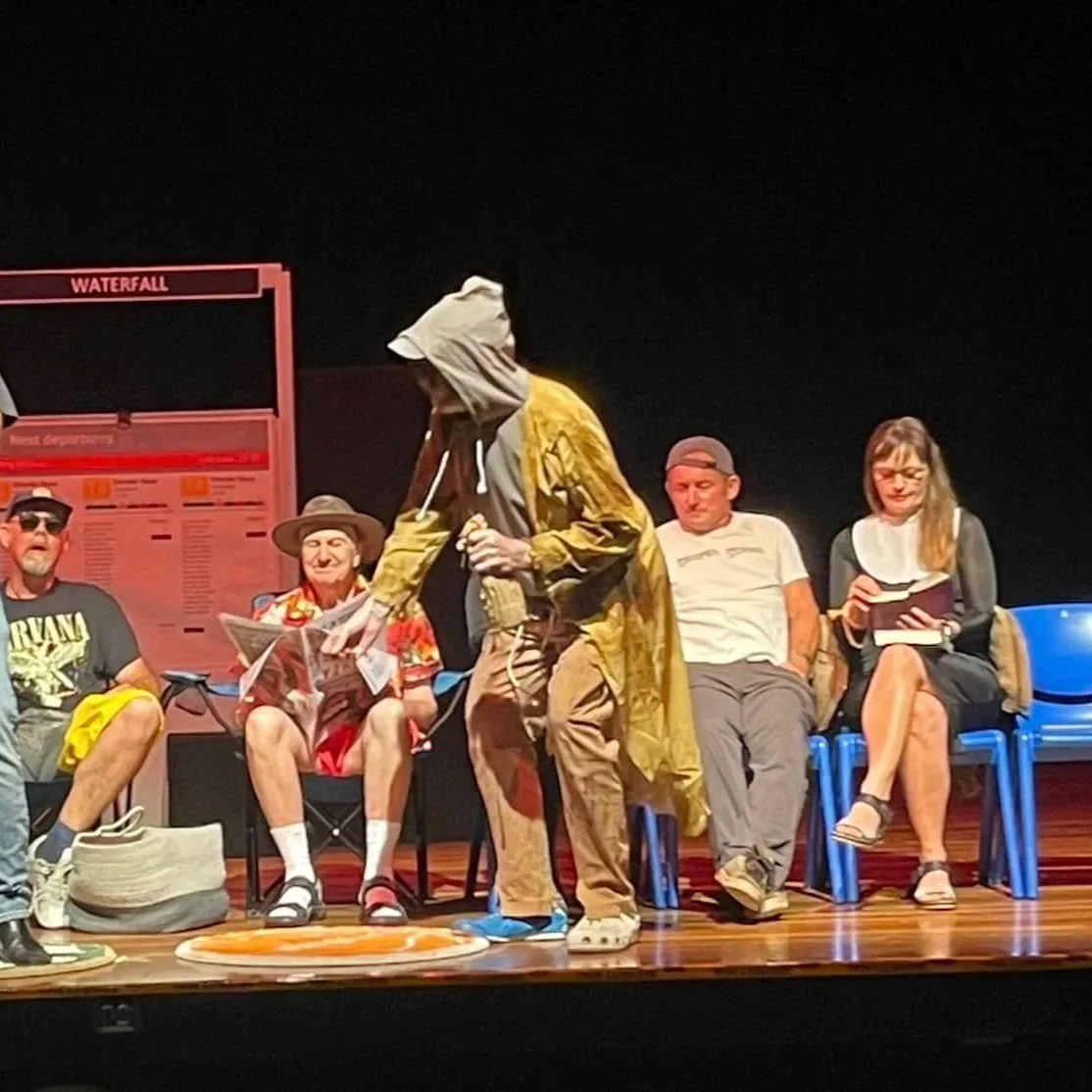 A group of five people sitting on chairs on a stage with a black background, with a mannequin dressed in traveler clothes, including a raincoat and a hat, standing in front of them.
