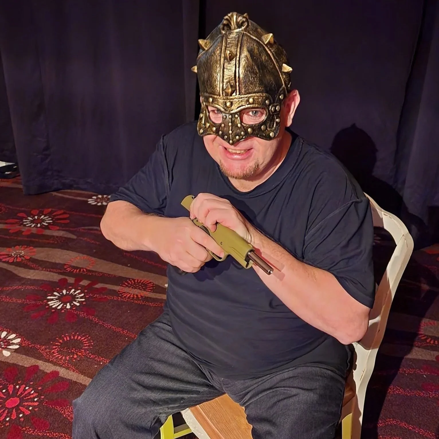 A man wearing a decorative Viking helmet and mask, sitting on a chair, holding a toy gun, smiling, on a patterned carpet with dark curtains in the background.