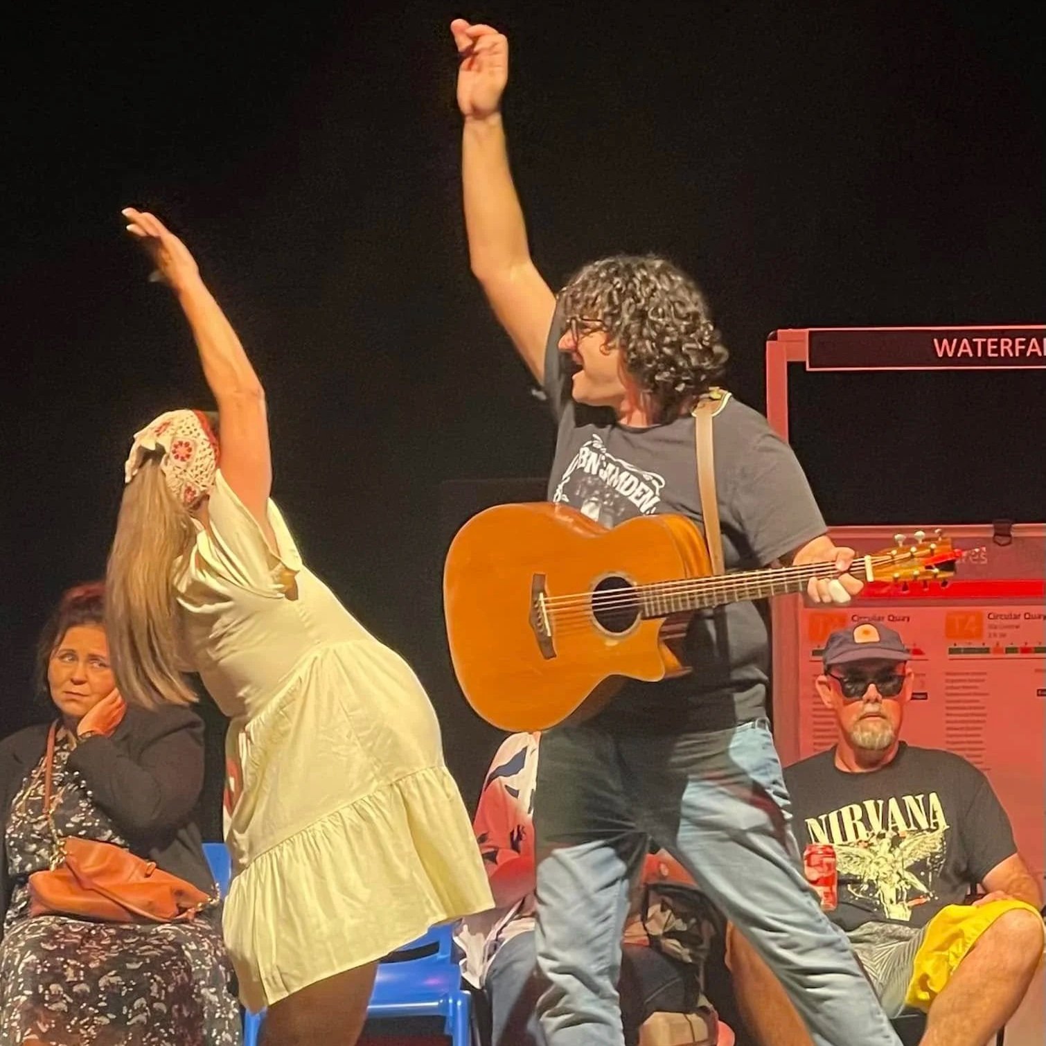 Two performers dancing on stage, one woman wearing a yellow dress with a headscarf, and one man playing a guitar, both raising their arms. In the background, two seated men, one with a Nirvana t-shirt and sunglasses.