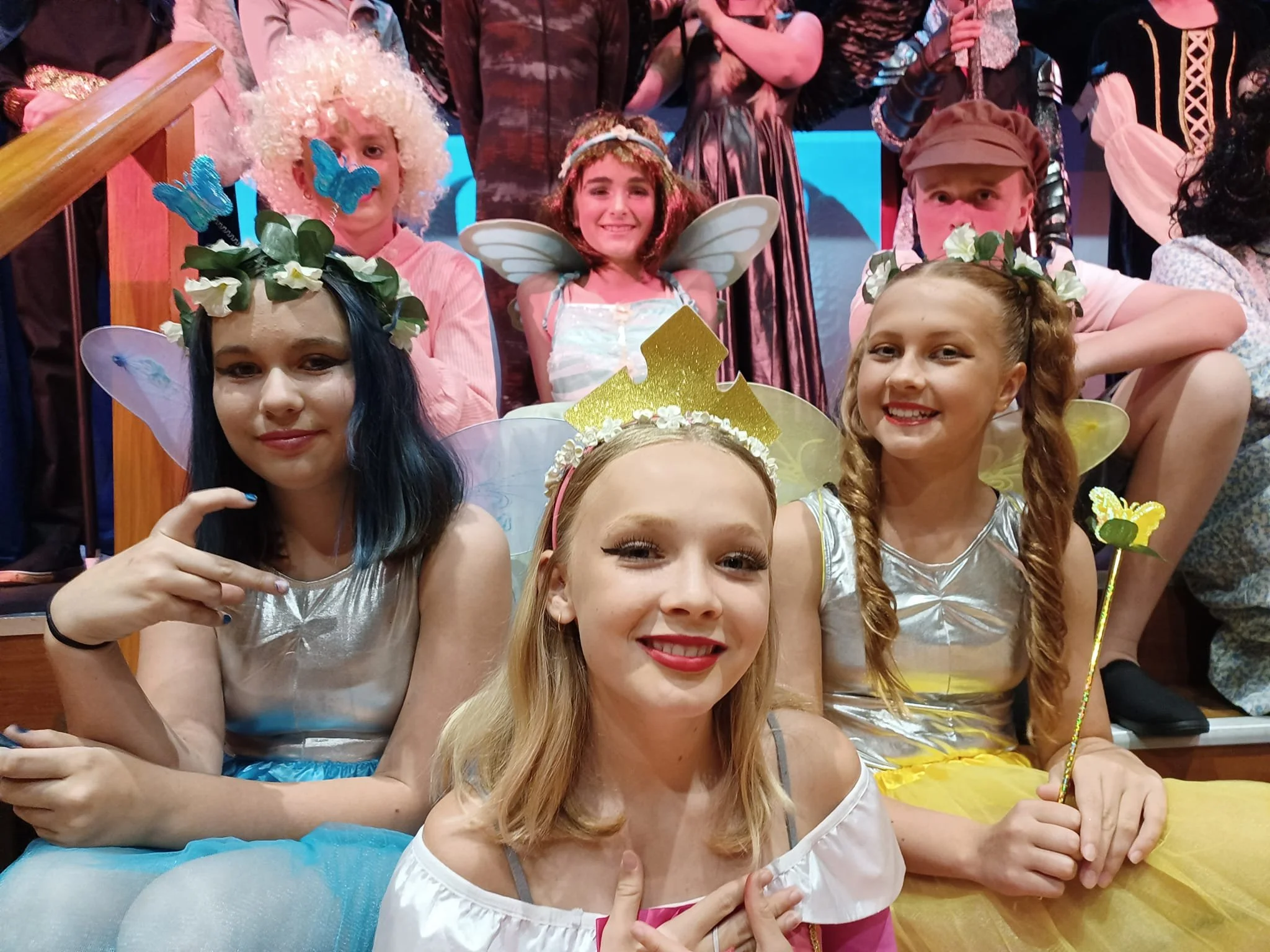 Group of children dressed as fairies with colorful wings and floral headbands, smiling at a costume event or performance.