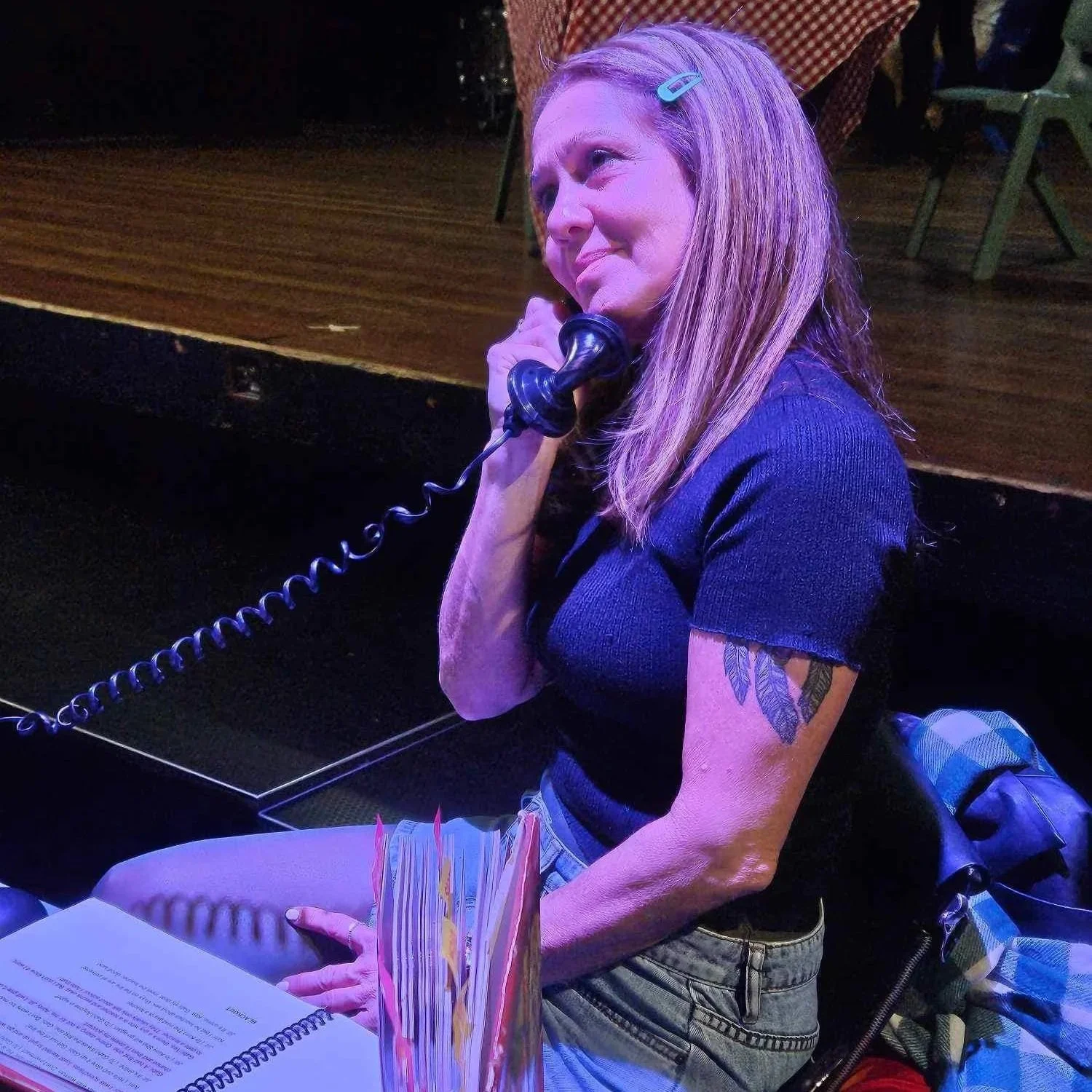 A woman with red hair, wearing a blue top, sitting on a stool with a phone receiver to her ear, looking to the side with a concerned expression. There is a notebook open in front of her and a colorful paper menu or fan in the foreground.