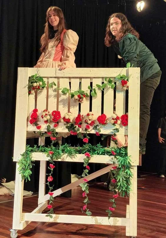 Two women standing behind a decorated wooden curtain on a stage, with one smiling and the other looking surprised.