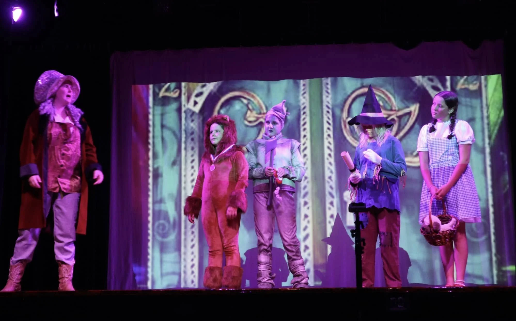 Children performing in a play on stage, dressed as fairy tale characters, with a colorful background projection.