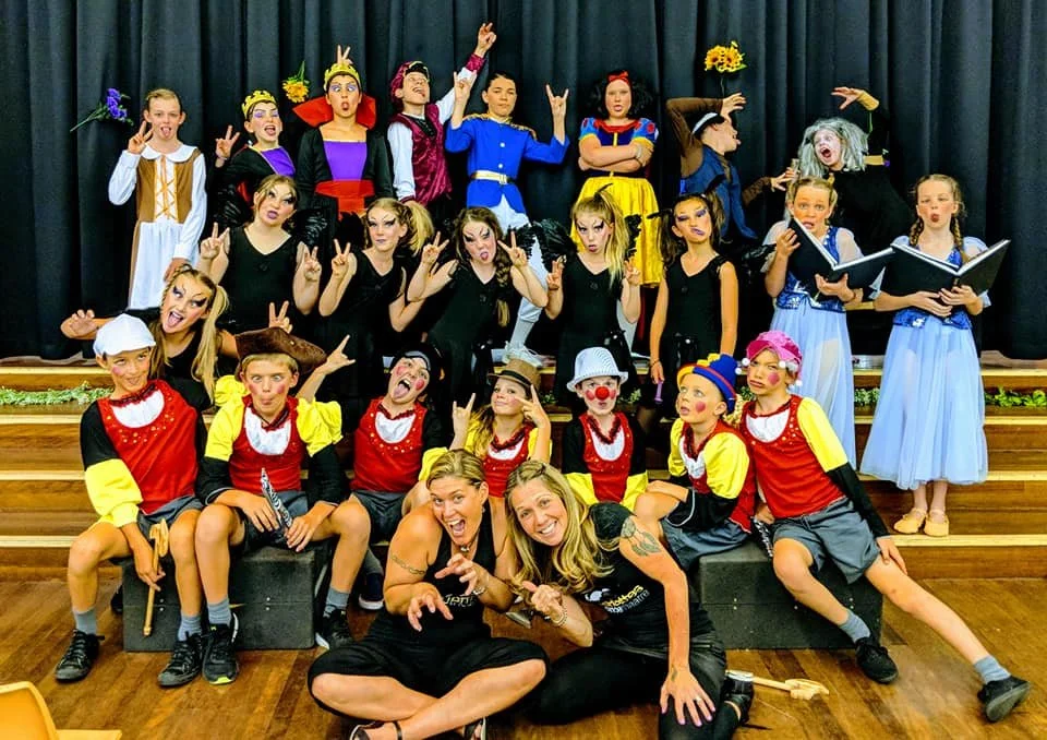 Children and adults dressed in colorful costumes performing on a stage with a black curtain backdrop.