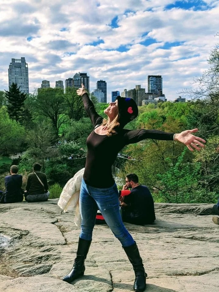 Woman posing with arms wide open on a rocky ledge in a park with trees, and city skyline in the background under a partly cloudy sky.