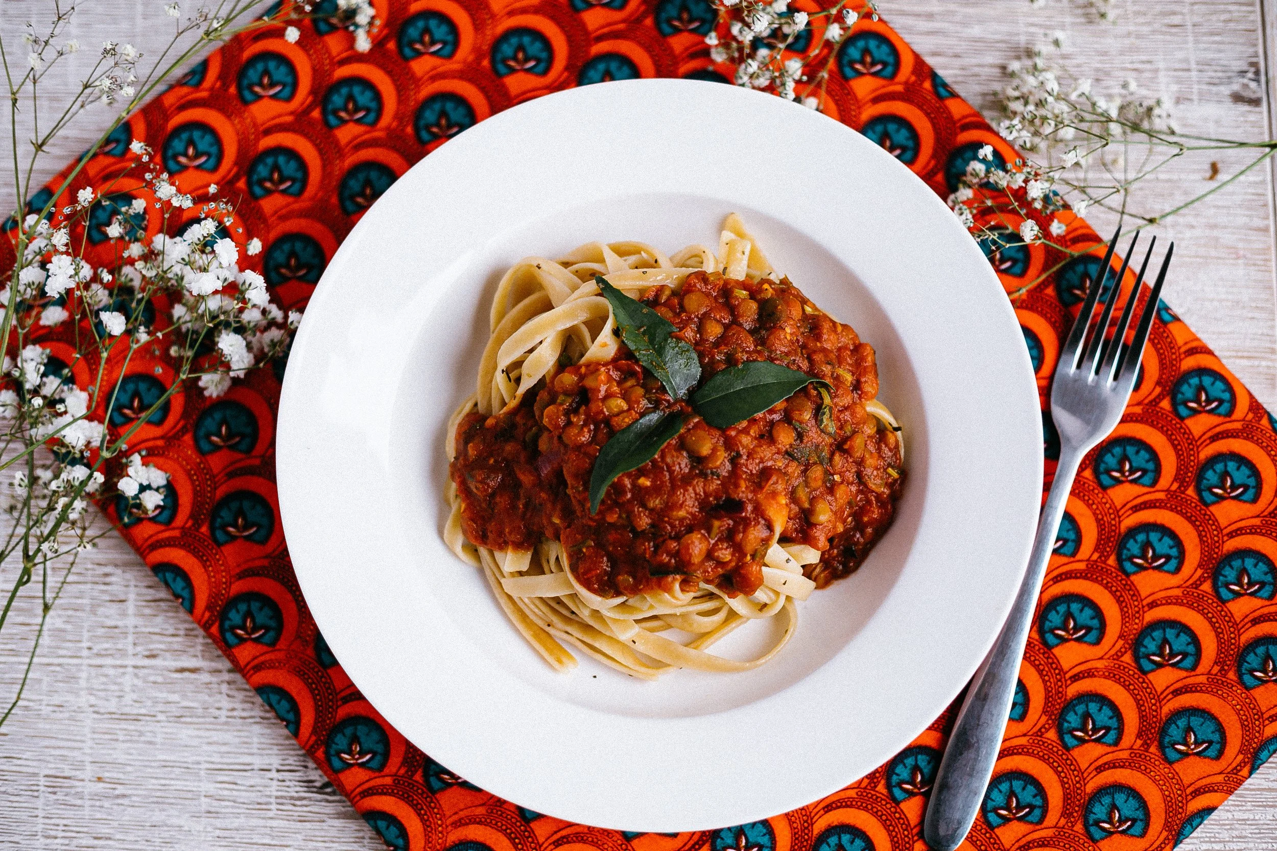 Plate of spaghetti with lentil tomato sauce garnished with bay leaves, served on white plate with a fork, set on a colorful patterned cloth with white flowers beside.