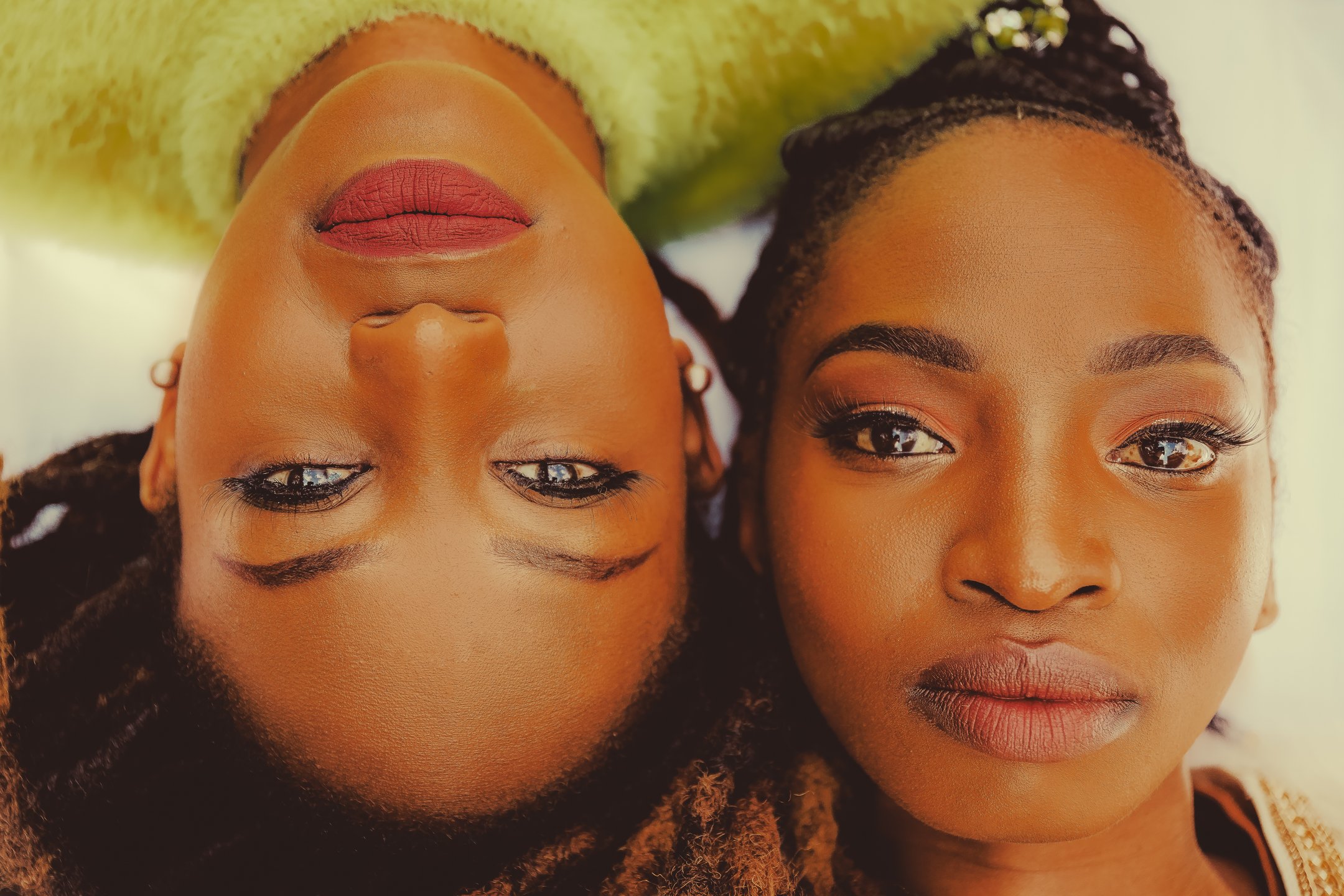 Close-up of two women lying down with their heads close together, one upside down, both looking directly at the camera. They have natural makeup, including eye makeup and lipstick, and braided hairstyles.