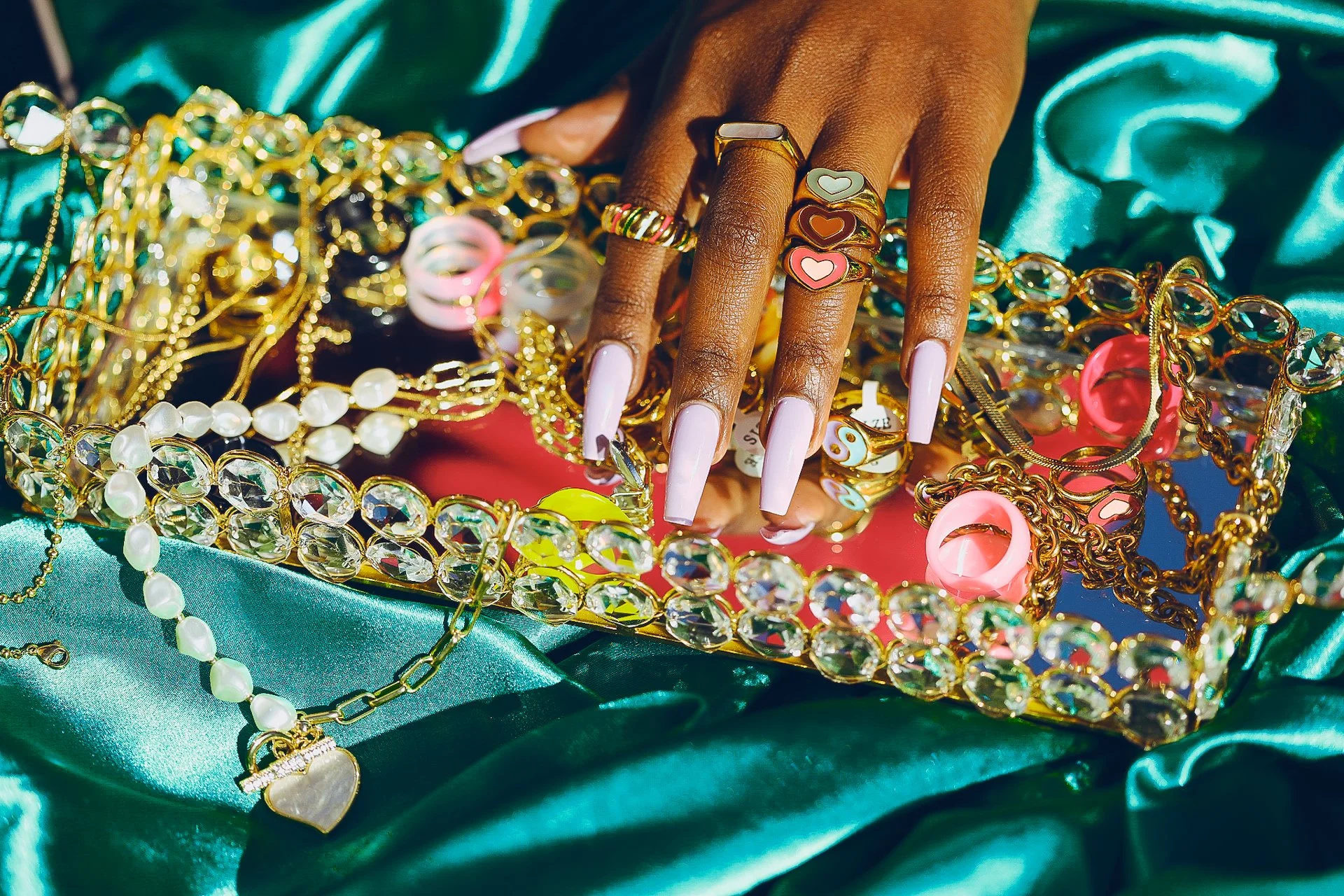 Hand with long, light pink nails resting on a tray of assorted jewelry, including gold and bejeweled necklaces, rings, and bracelets, placed on teal satin fabric.