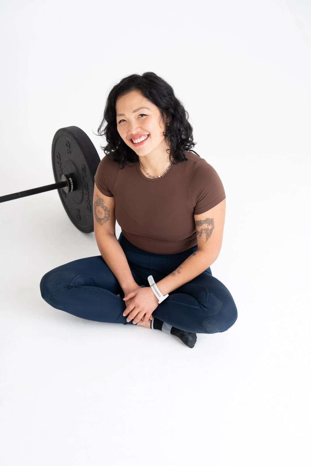 A young woman with long black hair, wearing a black camouflage tank top and a necklace, smiling in front of a white brick wall.