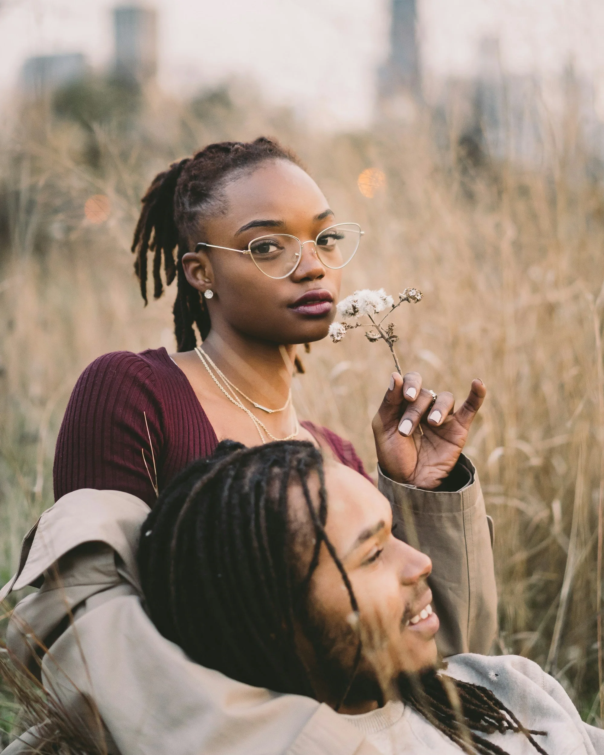 A woman with glasses and jewelry holding a small dried flower, standing in a field with tall grass, with buildings in the background. She has dreadlocks and is wearing a purple sweater.