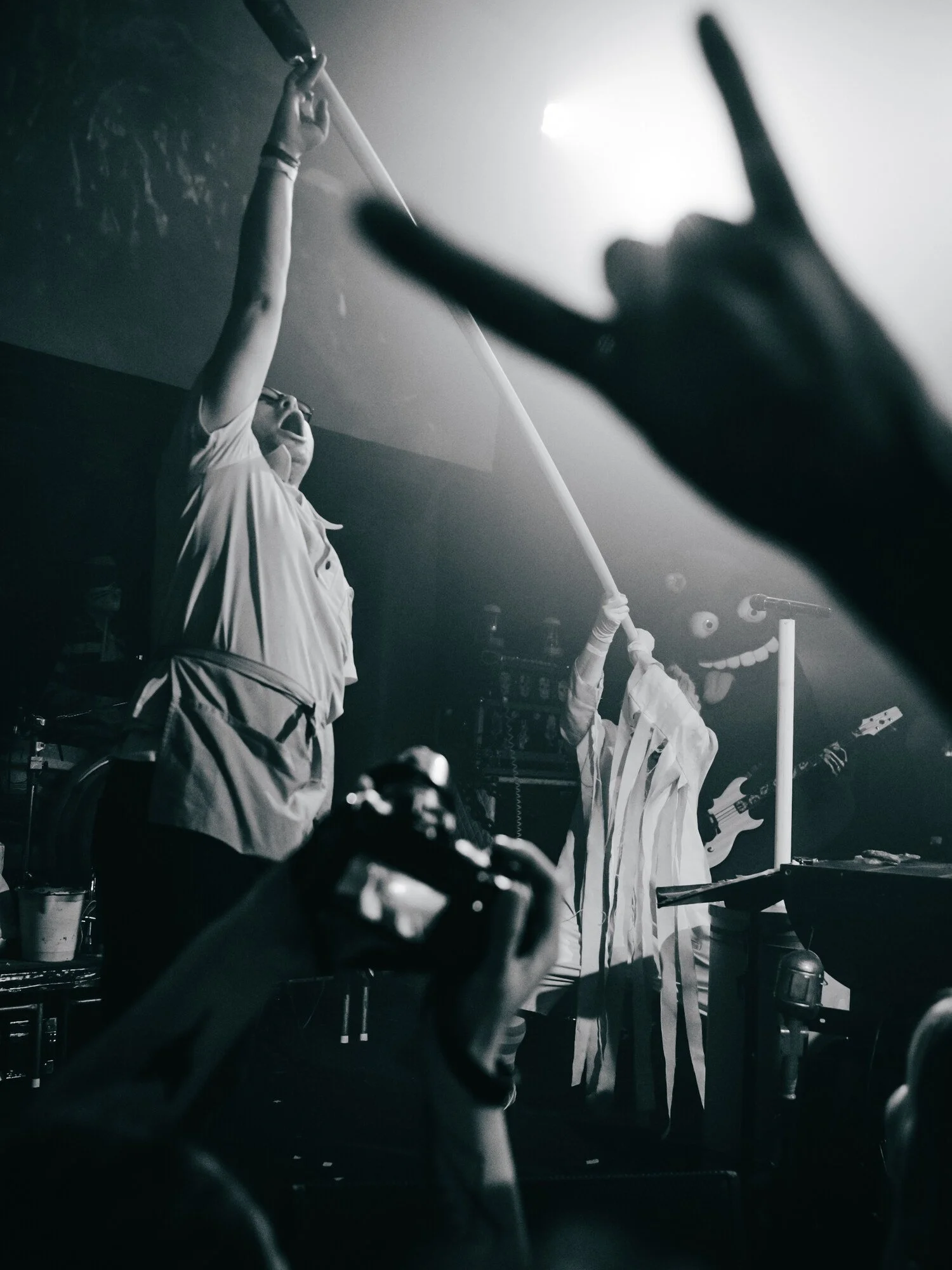 Two performers on stage, one holding a microphone stand upright, with energetic expressions; another with a guitar in the background; audience members with cameras capturing the moment. Black and white.