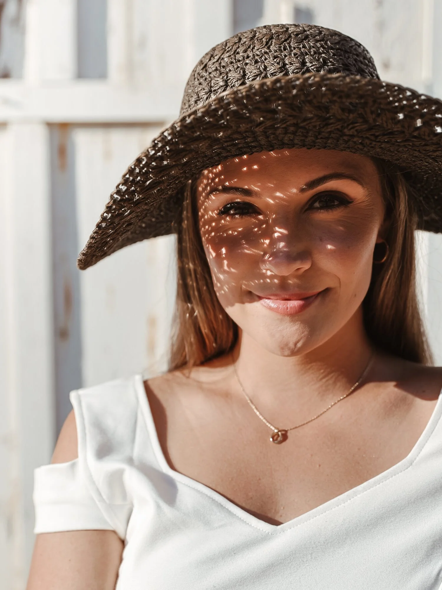 Smiling woman wearing a large woven sunhat, a white top with shoulder cut-outs, and a necklace, posing outdoors with sunlight creating patterns on her face.