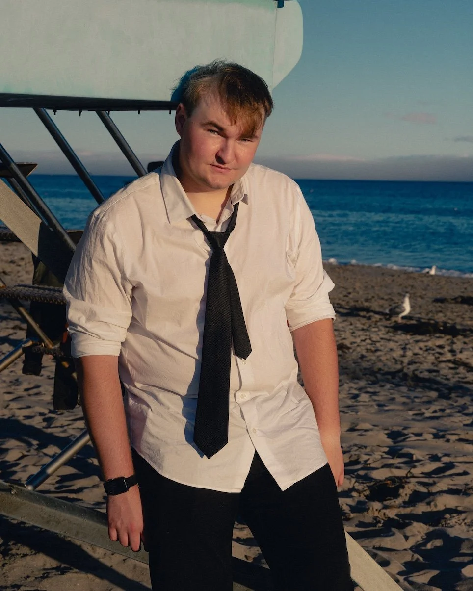 Young man wearing a white shirt and black tie standing on a beach near the ocean during sunset.